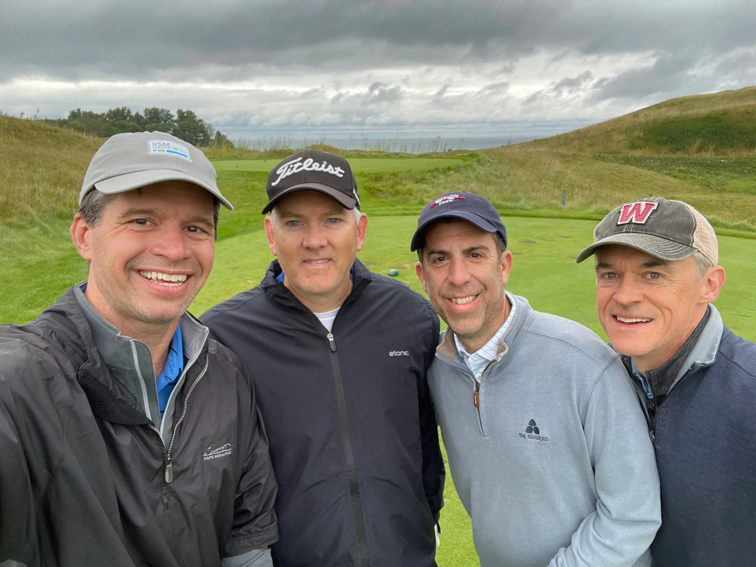 Four men in golf attire standing on a golf course with overcast sky and rolling hills in the background.