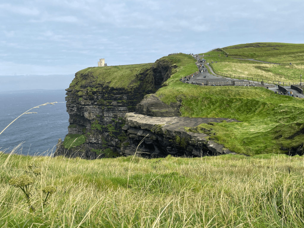 The Cliffs of Moher in Ireland with a grassy hill, stone staircase, and a small castle in the distance, overlooking the ocean