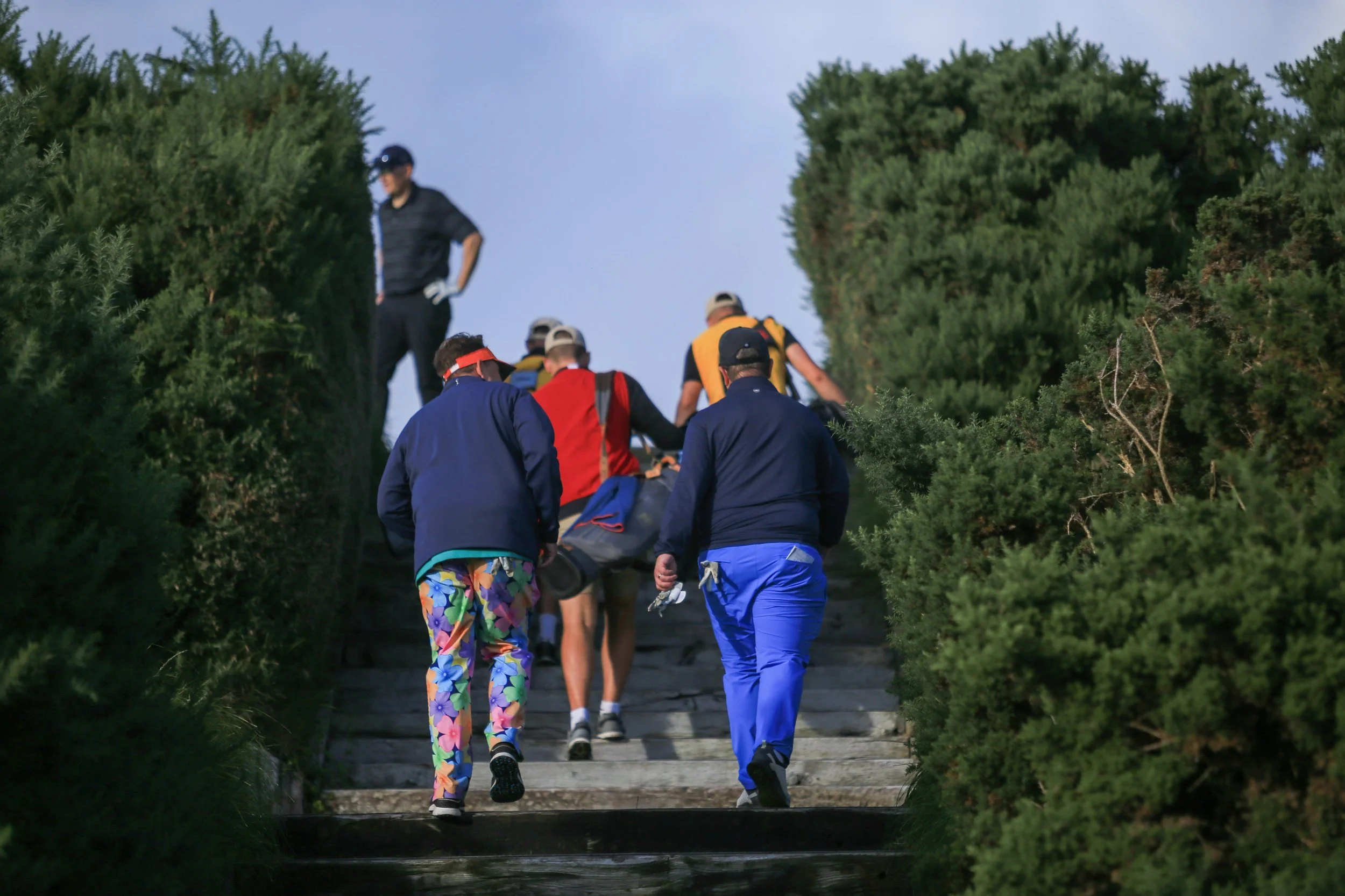 Group of people walking up outdoor steps surrounded by greenery, some wearing colorful clothing and carrying backpacks. Royal County Down, Northern Ireland. Caddies, buddies golf trip. Trip of a lifetime. Dream golf.