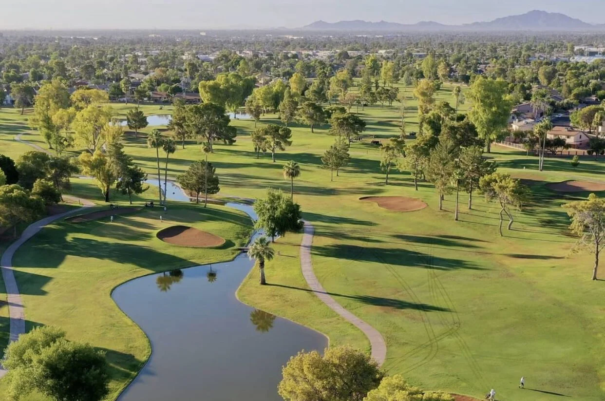 Aerial view of a golf course with green fairways, sand bunkers, water hazards, and scattered trees, with houses and mountains in the background.  Dobson Ranch Golf.  Mesa, Arizona.