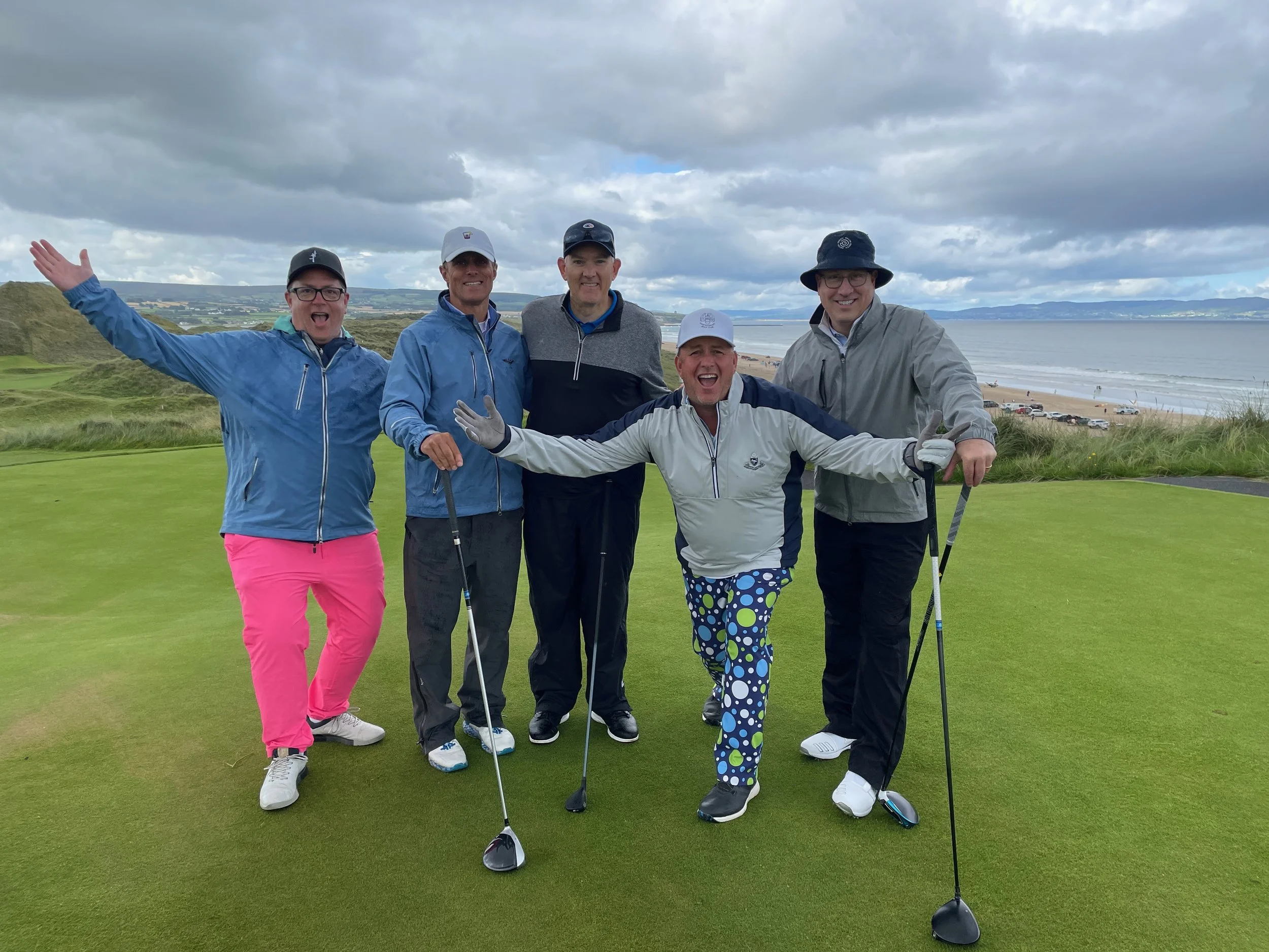 Five men celebrating on a golf course near the ocean, with cloudy skies overhead.