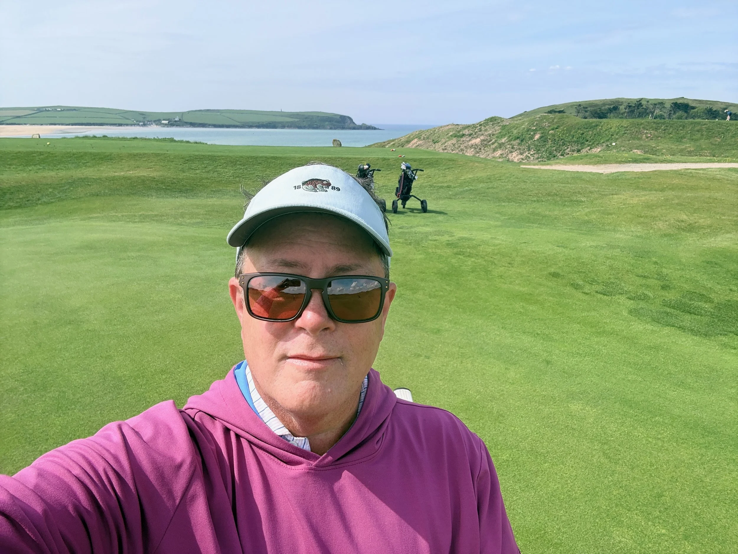 Founder and CEO Matt Greber wearing sunglasses, a white visor with a logo, and a purple jacket is taking a selfie on a golf course with green grass, a pathway, and a view of water and rolling hills in the background.