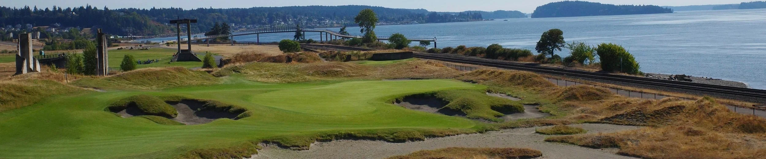 A coastal golf course with green grass, sand traps, and rolling hills, overlooking water with bridges and trees in the background.