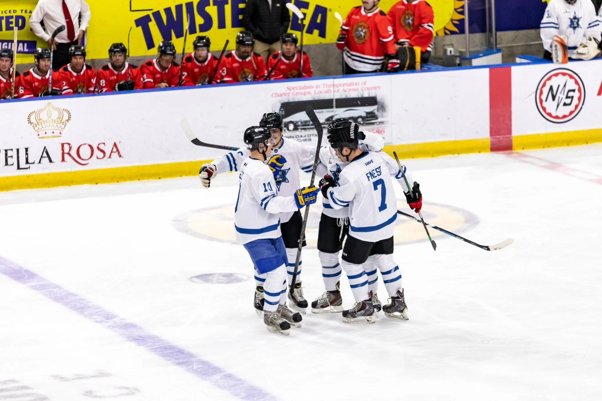 Ice hockey players in white jerseys celebrating on the ice while other players in red jerseys sit on the bench in the background.