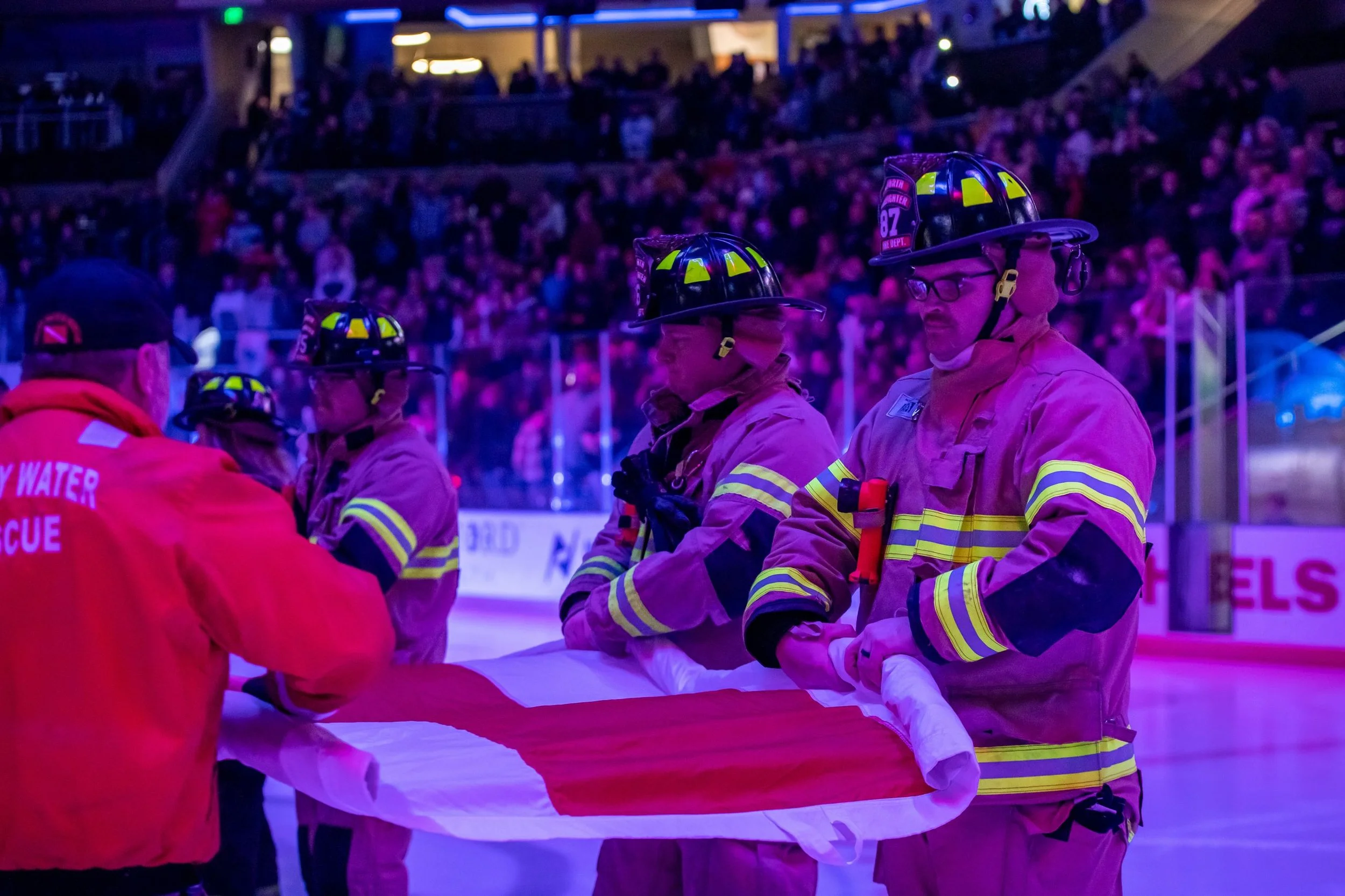 Firefighters in uniform and helmets are working together to handle an emergency. They are inside a building with a crowd of people seated in the background, under blue and purple lighting.