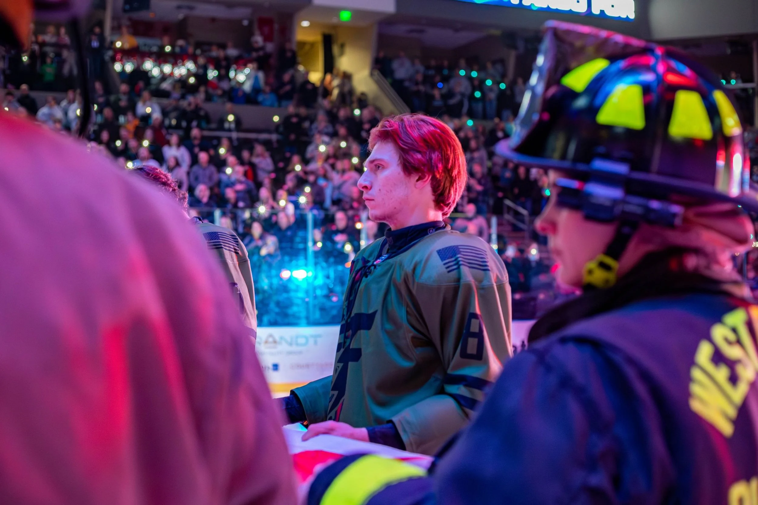 Two security personnel wearing uniforms and helmets standing in an indoor sports arena filled with spectators, with bright lights and electronic displays in the background.