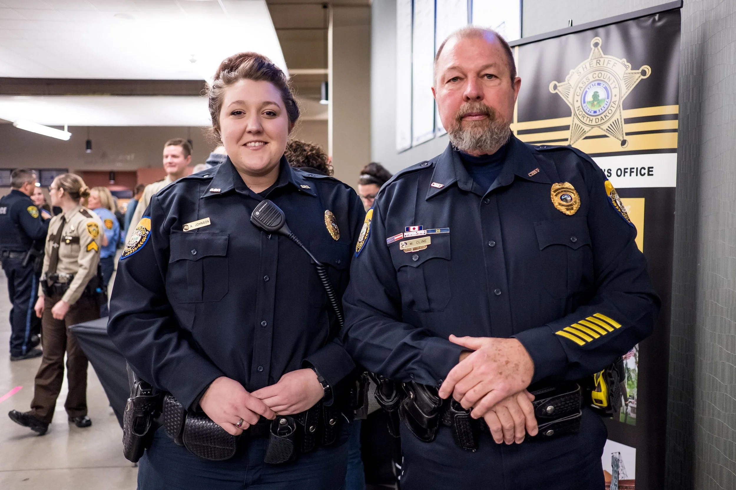 Two police officers, a female and a male, standing side by side inside a building, smiling at the camera. There are other officers and people in the background, and a sheriff's badge logo on a banner behind them.