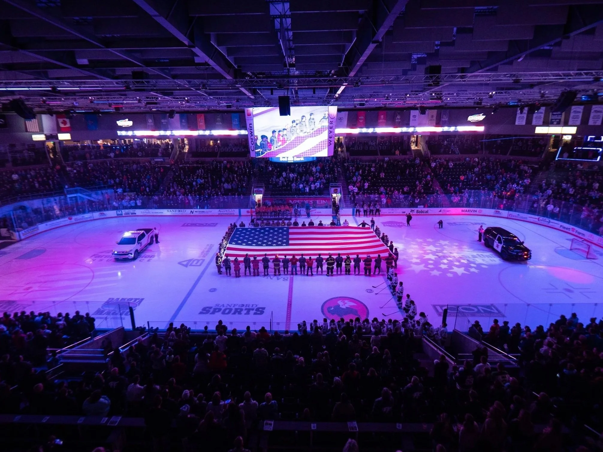 An ice hockey arena filled with spectators, with a large American flag ice rink at the center and players lined up for the national anthem before a game.