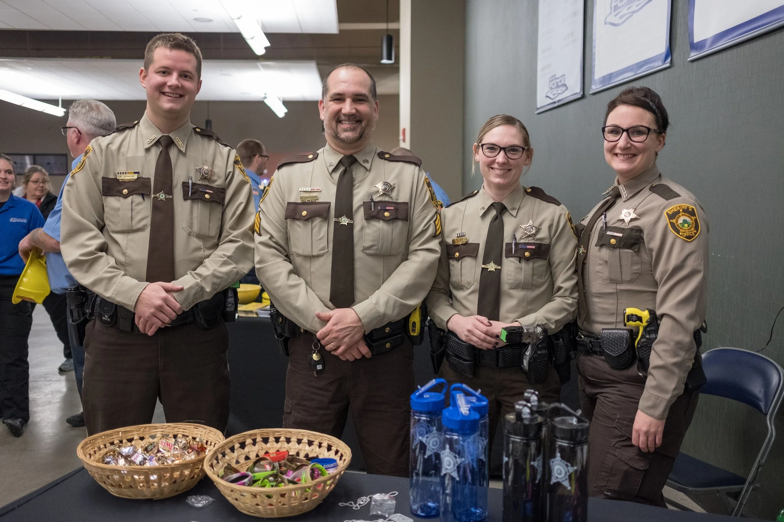 Group of four law enforcement officers smiling and standing at a table with baskets of candy and water bottles, in what appears to be an indoor community event or fair.
