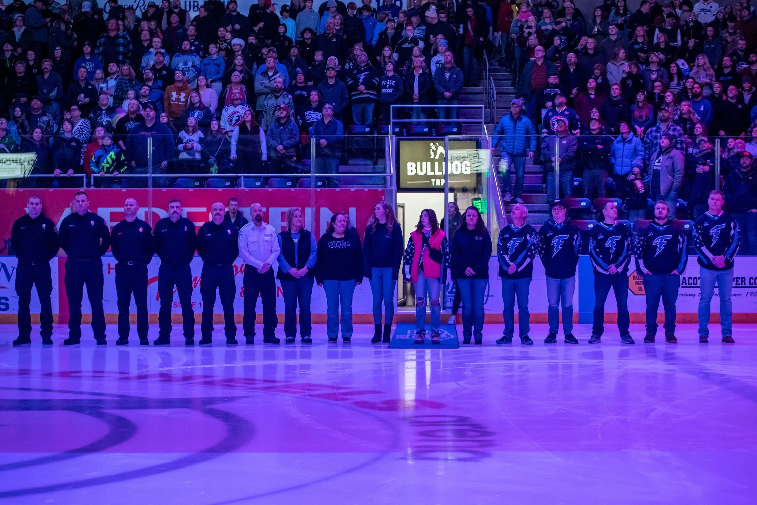 Ice hockey arena with players standing on the ice rink, audience in the stands, and officials lined up in front, during the national anthem or a moment of silence.