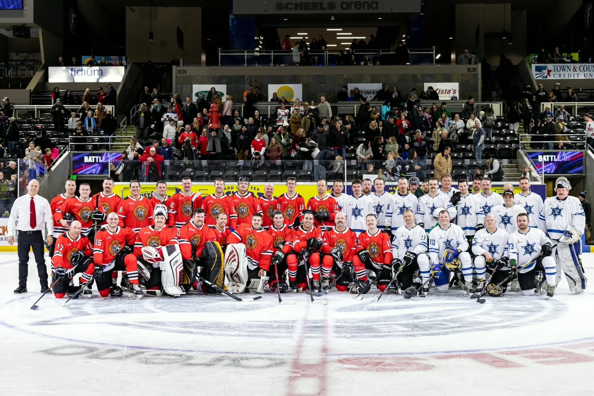 A group of hockey players in red and white jerseys posing on the ice with coaches and officials, in an indoor arena with spectators in the background.