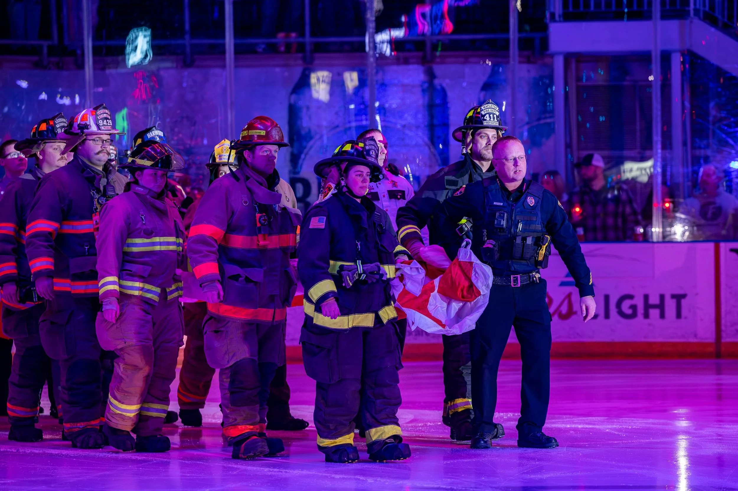 A group of firefighters in uniform standing on an ice rink, some with helmets, under colorful lighting.