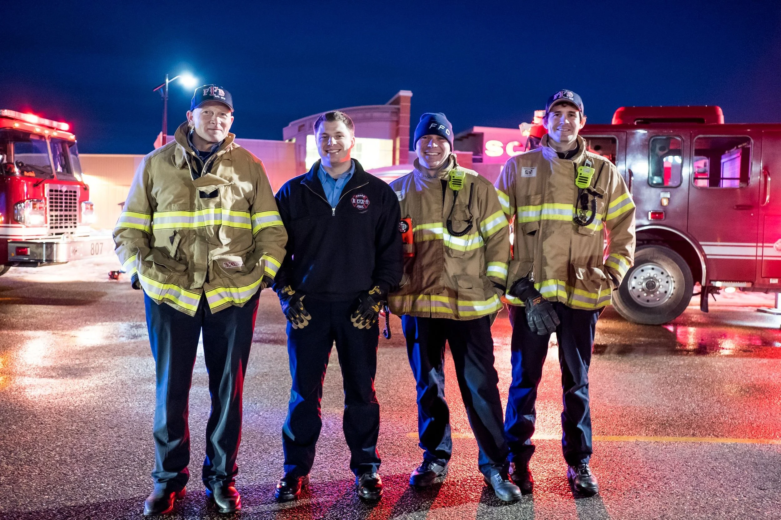 Four firefighters in their gear standing in front of fire trucks at night, smiling.