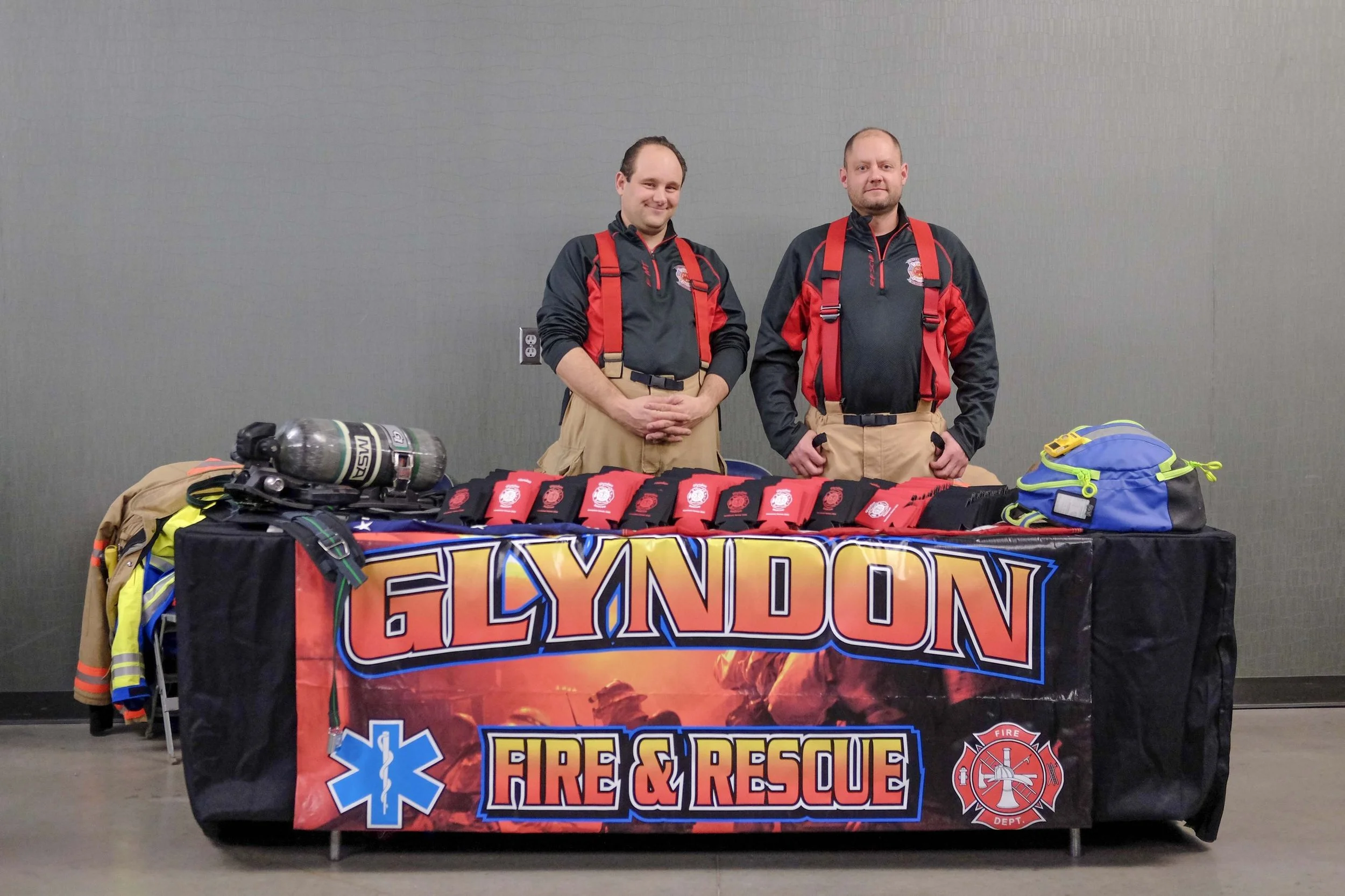 Two firefighters in uniform standing behind a table with firefighting gear, informational materials, and a banner reading 'Fire & Rescue.'