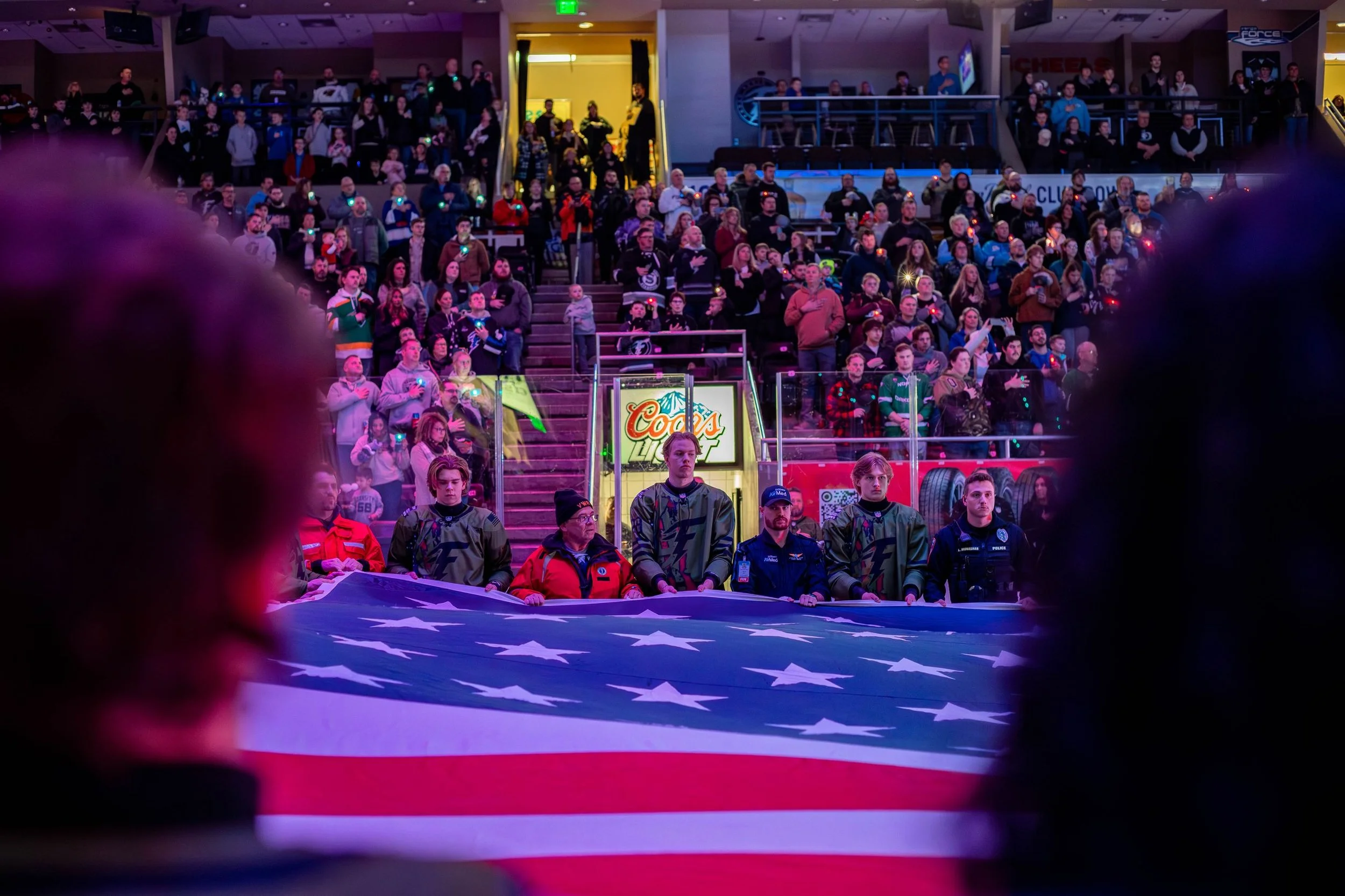 Crowd of people standing in an indoor arena, some with their hands over their hearts, with a large American flag being displayed in front of them. Spectators are on multiple levels, and some appear to be watching a ceremony or event.