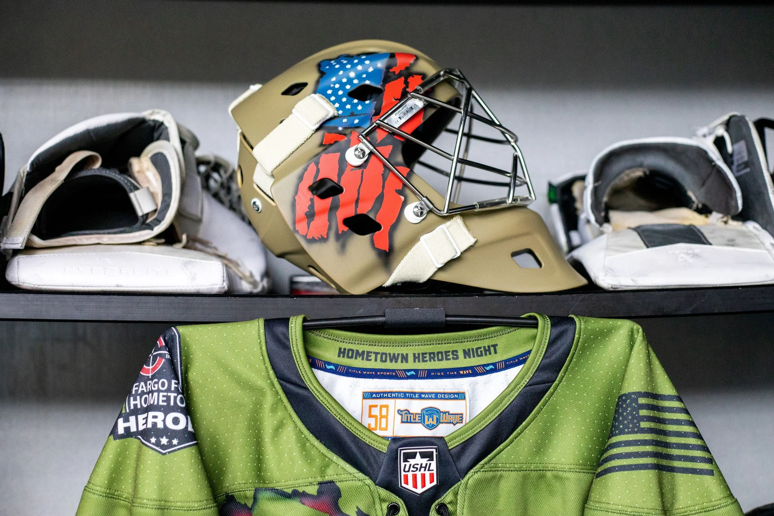 A hockey helmet with American flag and claw marks designs, hockey gloves on either side, and a green hockey jersey with various patches and logos underneath.