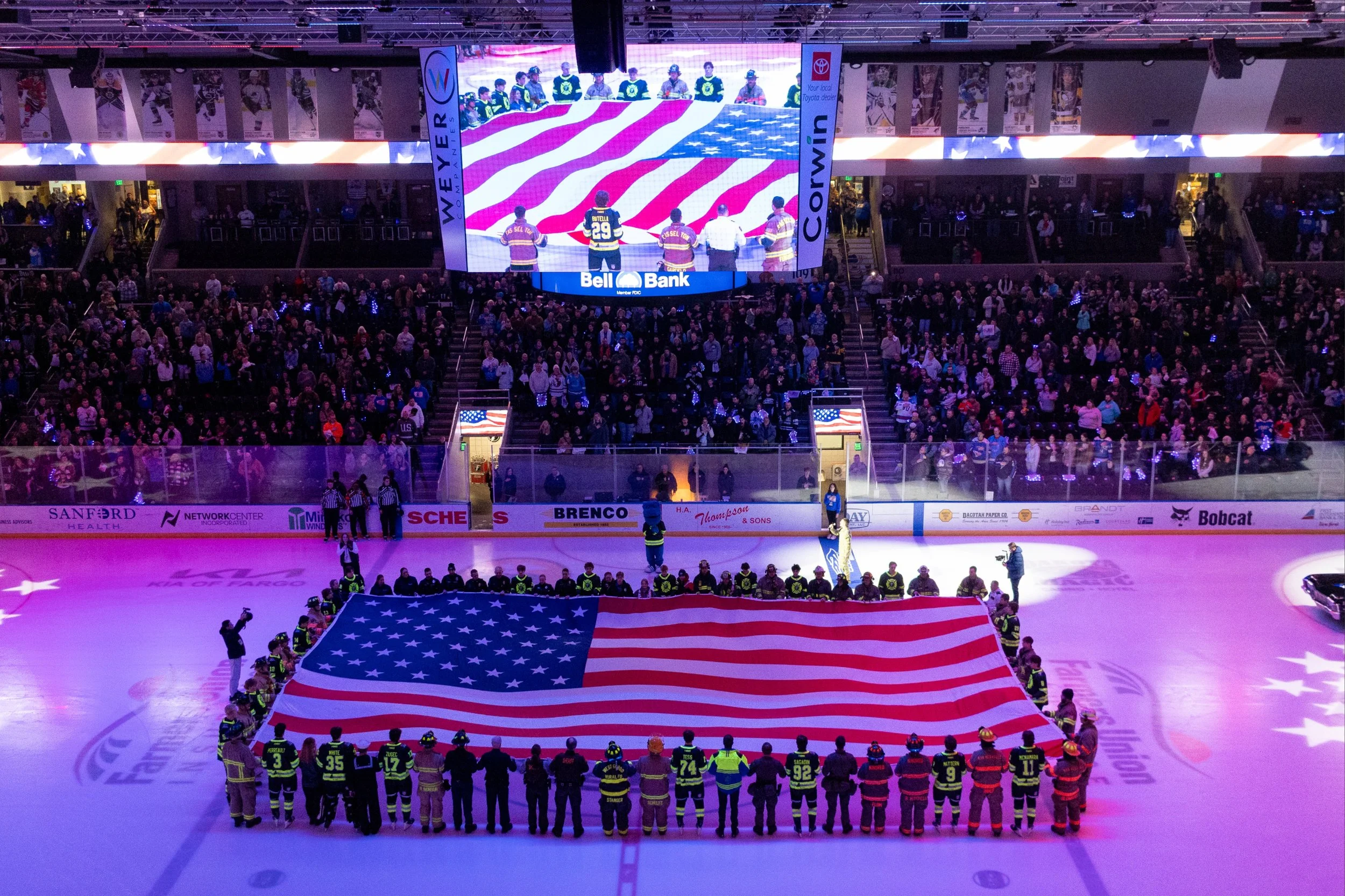 An indoor ice hockey rink filled with spectators. A large American flag is being held by players and team staff on the ice. The crowd is gathered in the stands, with some looking towards a big screen displaying players holding the American flag. The arena is decorated with patriotic elements.