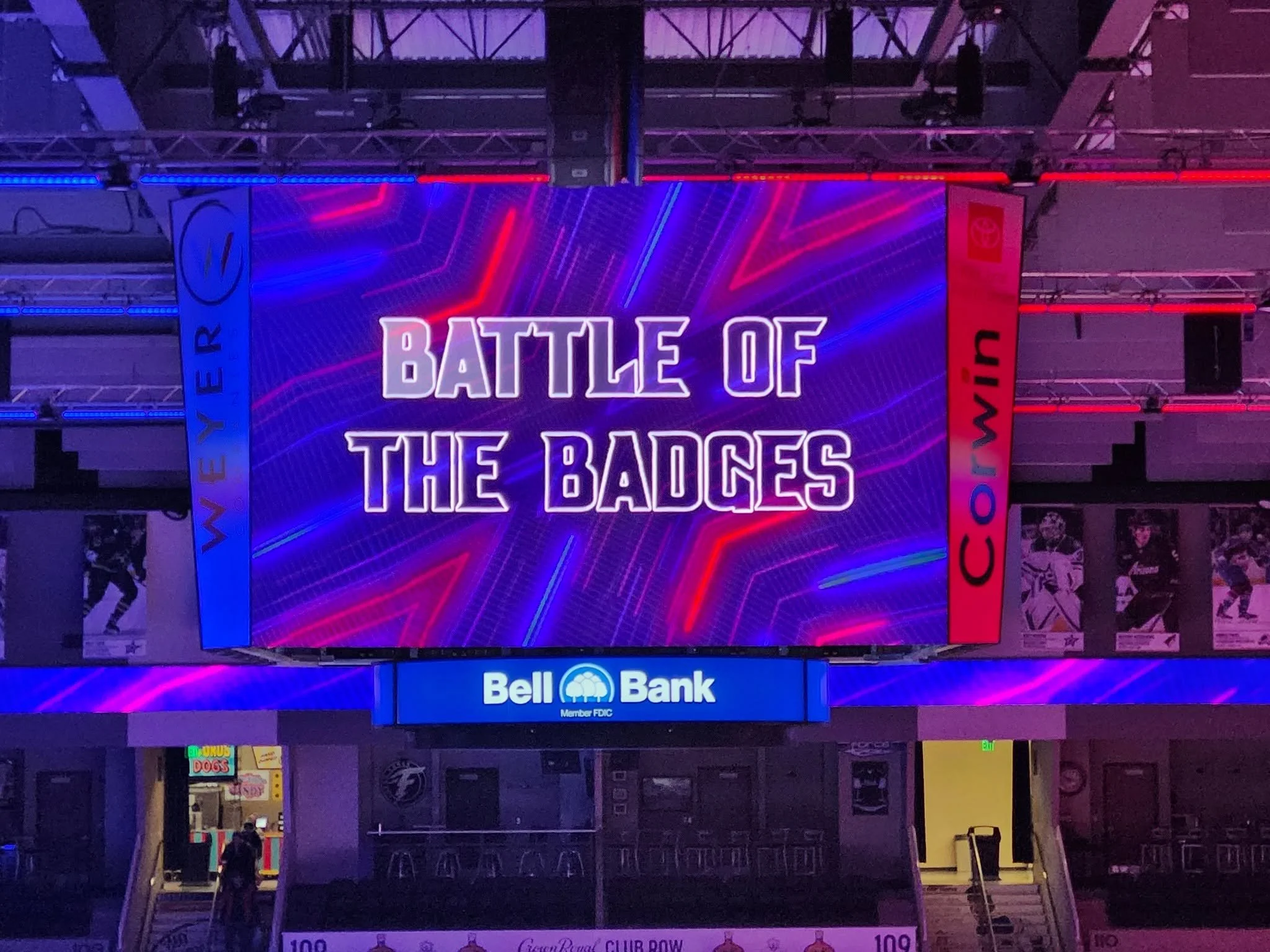 Bright neon sign reads 'Battle of the Bagues' in purple and pink colors. Surrounding structure has red and blue lighting, and there are billboards and posters visible below.