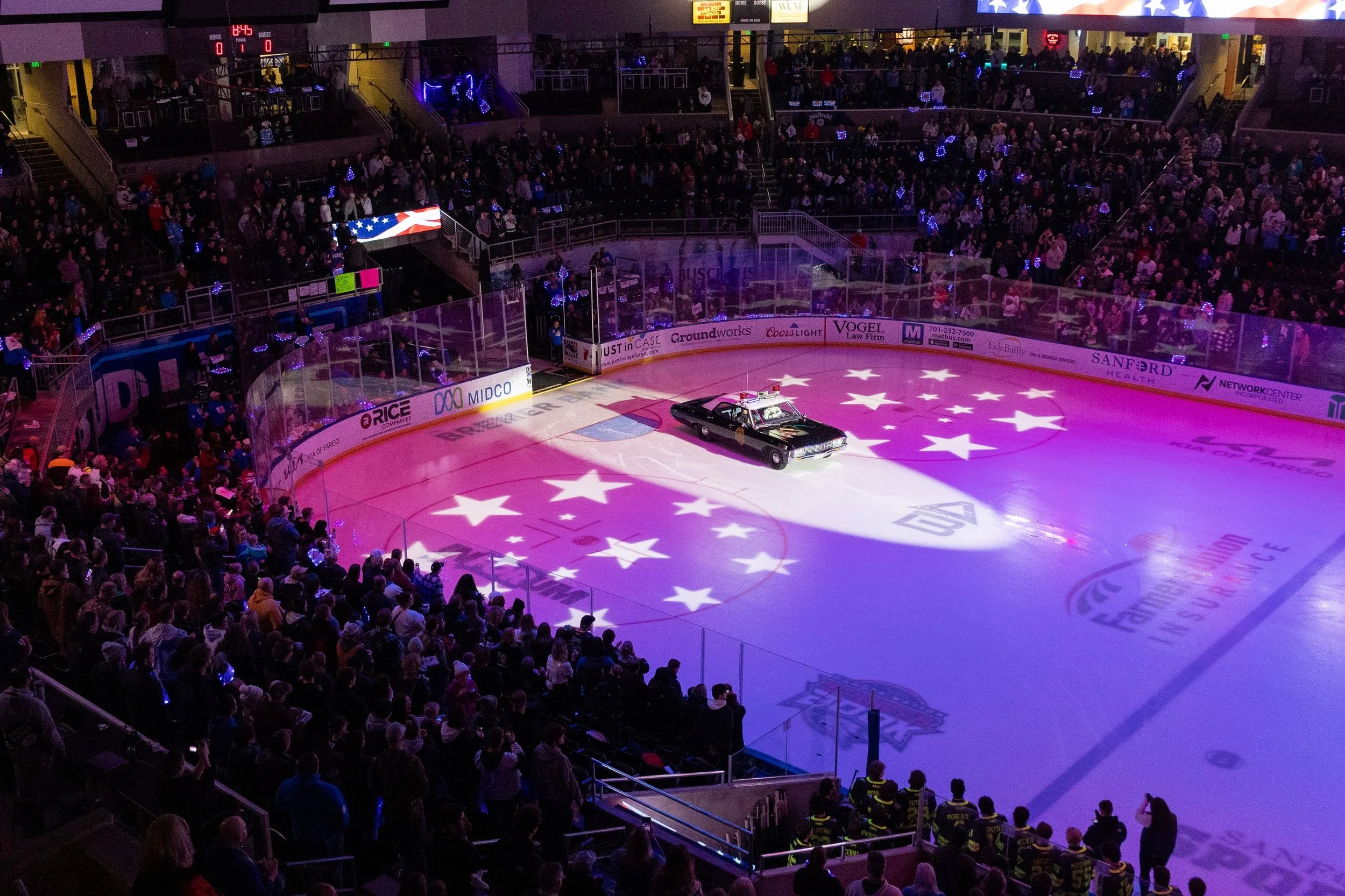 An indoor hockey arena filled with spectators, with a shining police car on the ice rink illuminated by stage lights that project the American flag. The arena is crowded with fans, and there are advertisements along the boards.