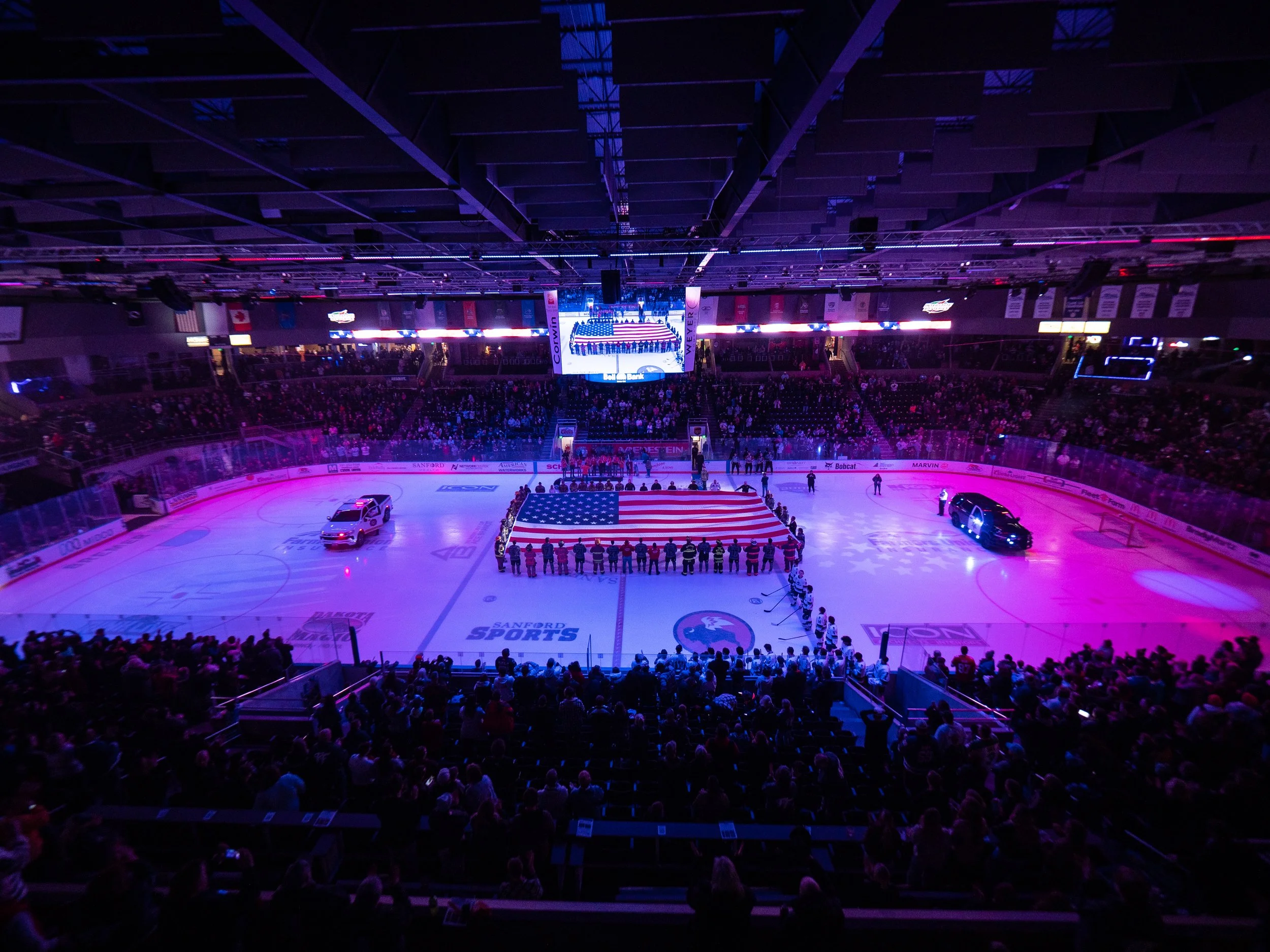 Indoor ice hockey arena with spectators, a large American flag covering the ice rink, and the national anthem being played.