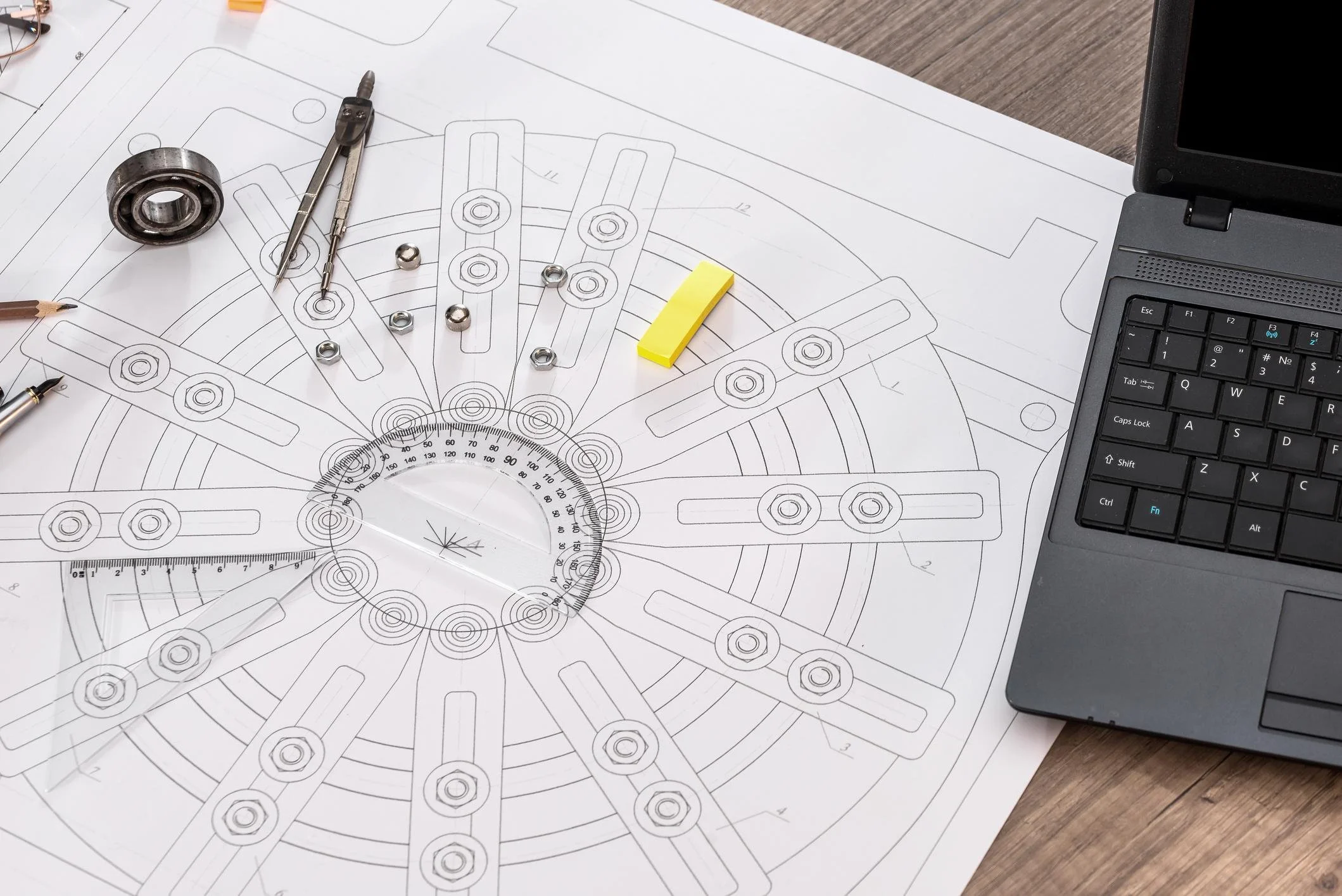 Technical drawing or blueprint of a mechanical part with various nuts, bolts, and tools placed on top. A yellow sticky note and part of a laptop keyboard are also visible.