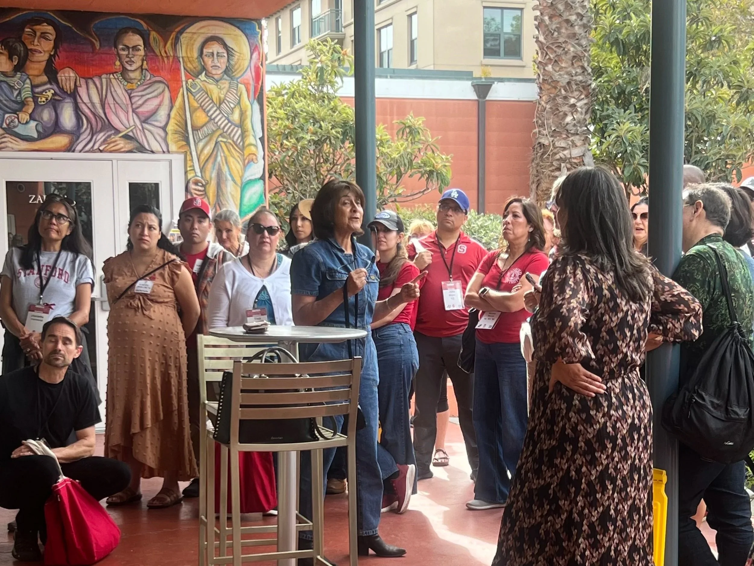 A diverse group of people gathered outdoors, listening to a woman in a blue outfit speak. The scene includes campus buildings and a colorful mural that includes Frida Kahlo and other women in the background.