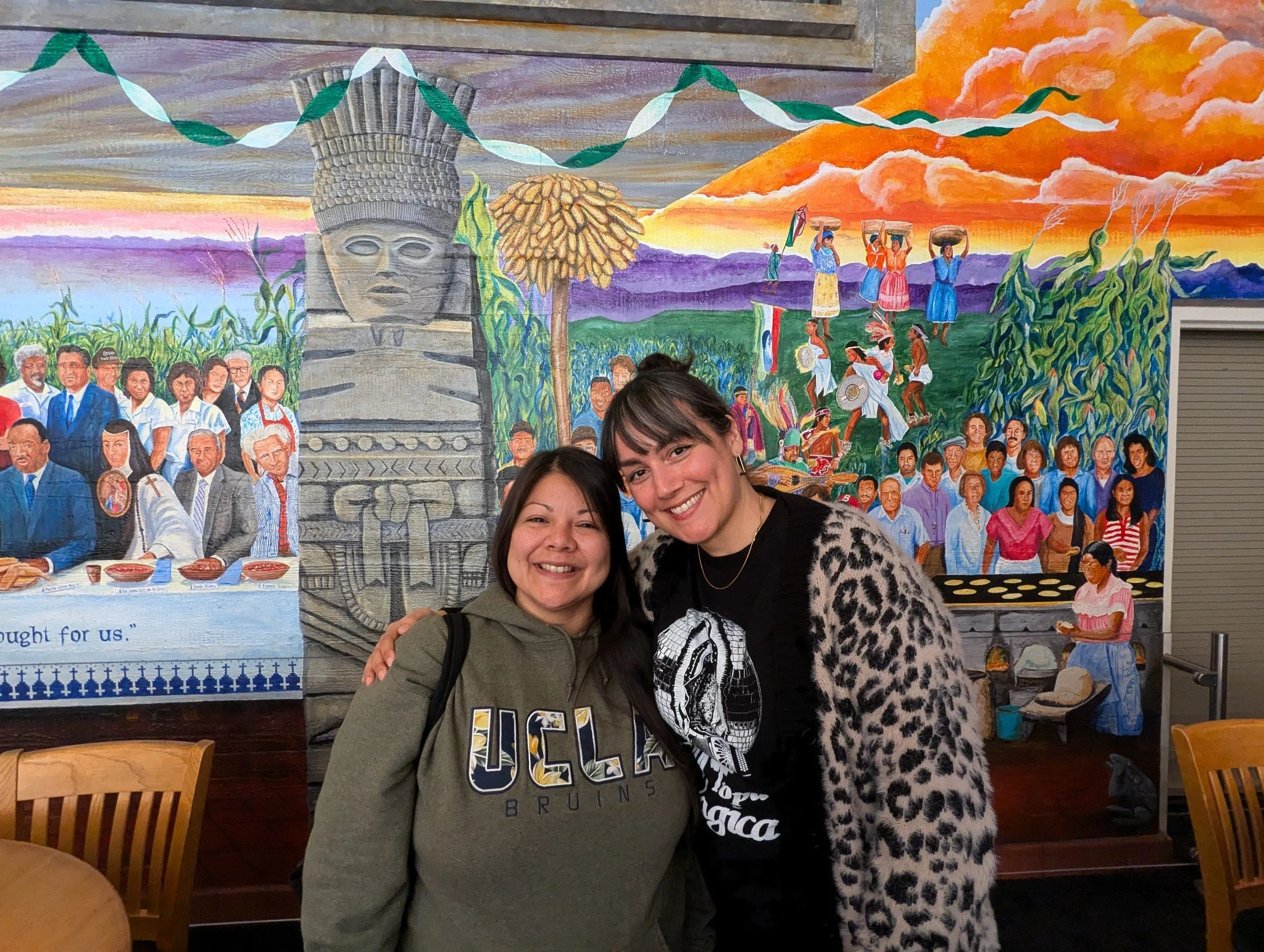 Two women smiling in front of a colorful mural depicting historical figures, community, and cultural scenes with a sunset sky in the background.