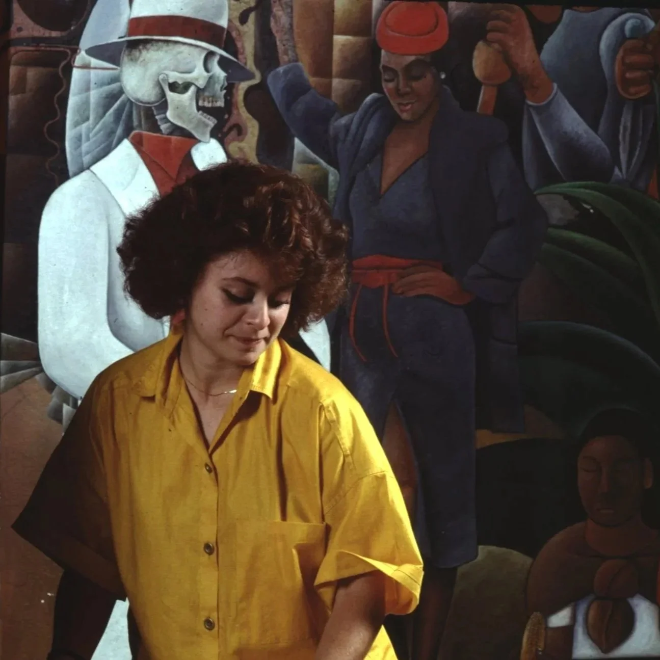 A woman with curly brown hair wearing a yellow shirt stands in front of a colorful mural featuring various characters, including a skeleton dressed in a Zoot Suit, a woman in a red hat, and other abstract figures.