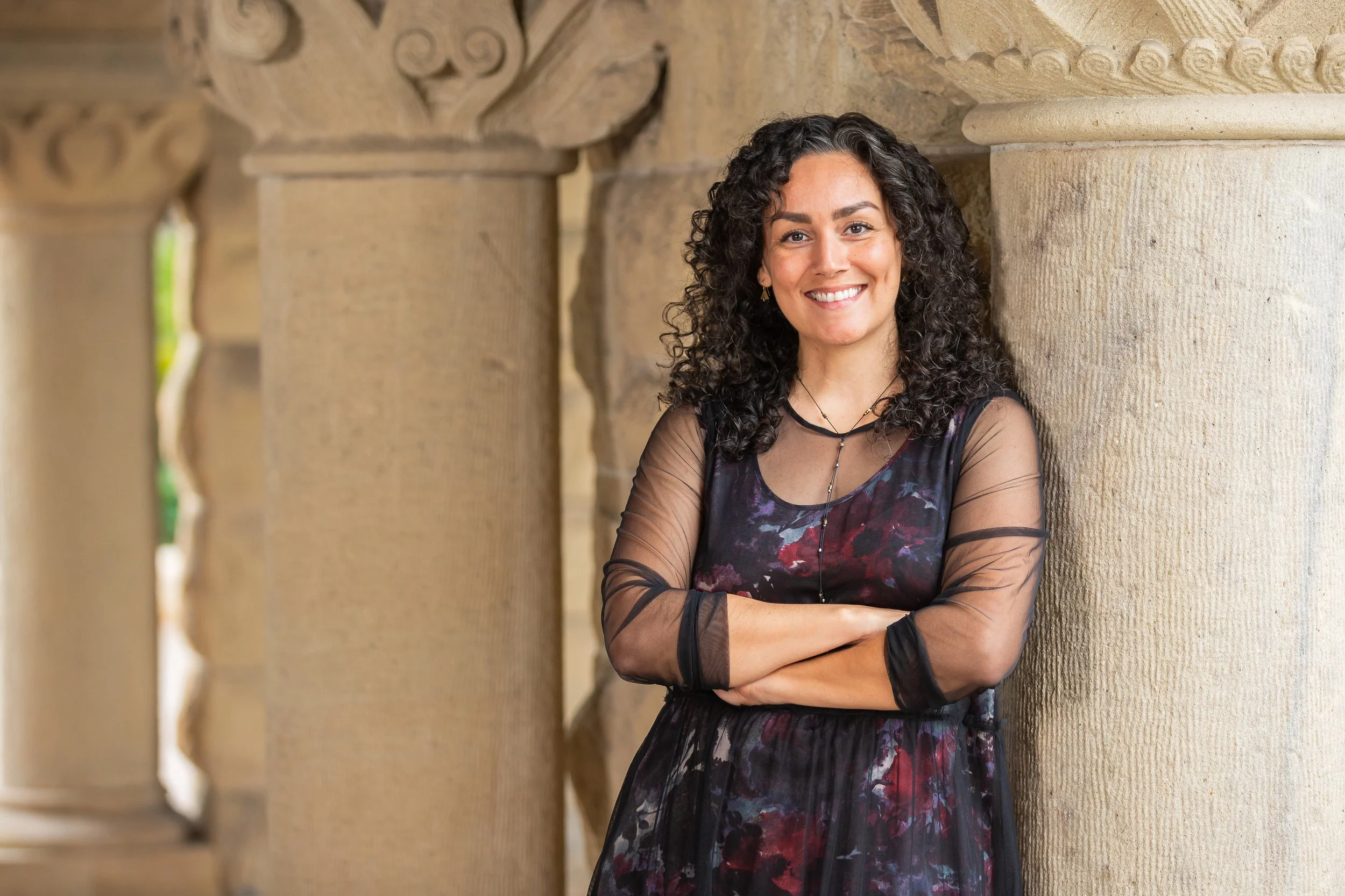 A woman with curly dark hair, wearing a black and purple floral dress with sheer black sleeves, leaning against a stone column with intricate carvings, smiling at the camera.