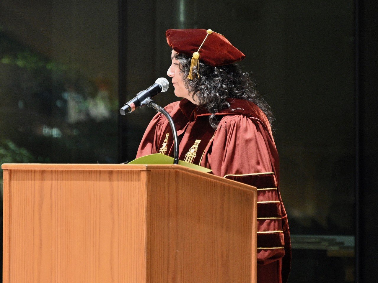 Rose Salseda wearing long curly hair and burnt orange colored doctoral regalia while speaking at a podium