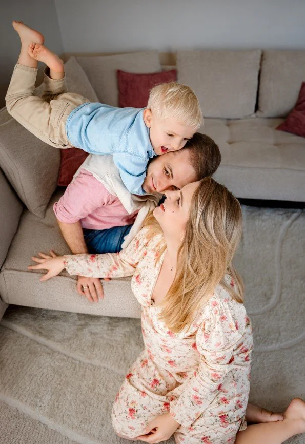 A woman with long blonde hair and a floral dress, a man with short brown hair wearing a pink and white shirt, and a young boy with blonde hair in a blue shirt and beige pants, playfully close together in a living room with a gray sofa and red cushions.