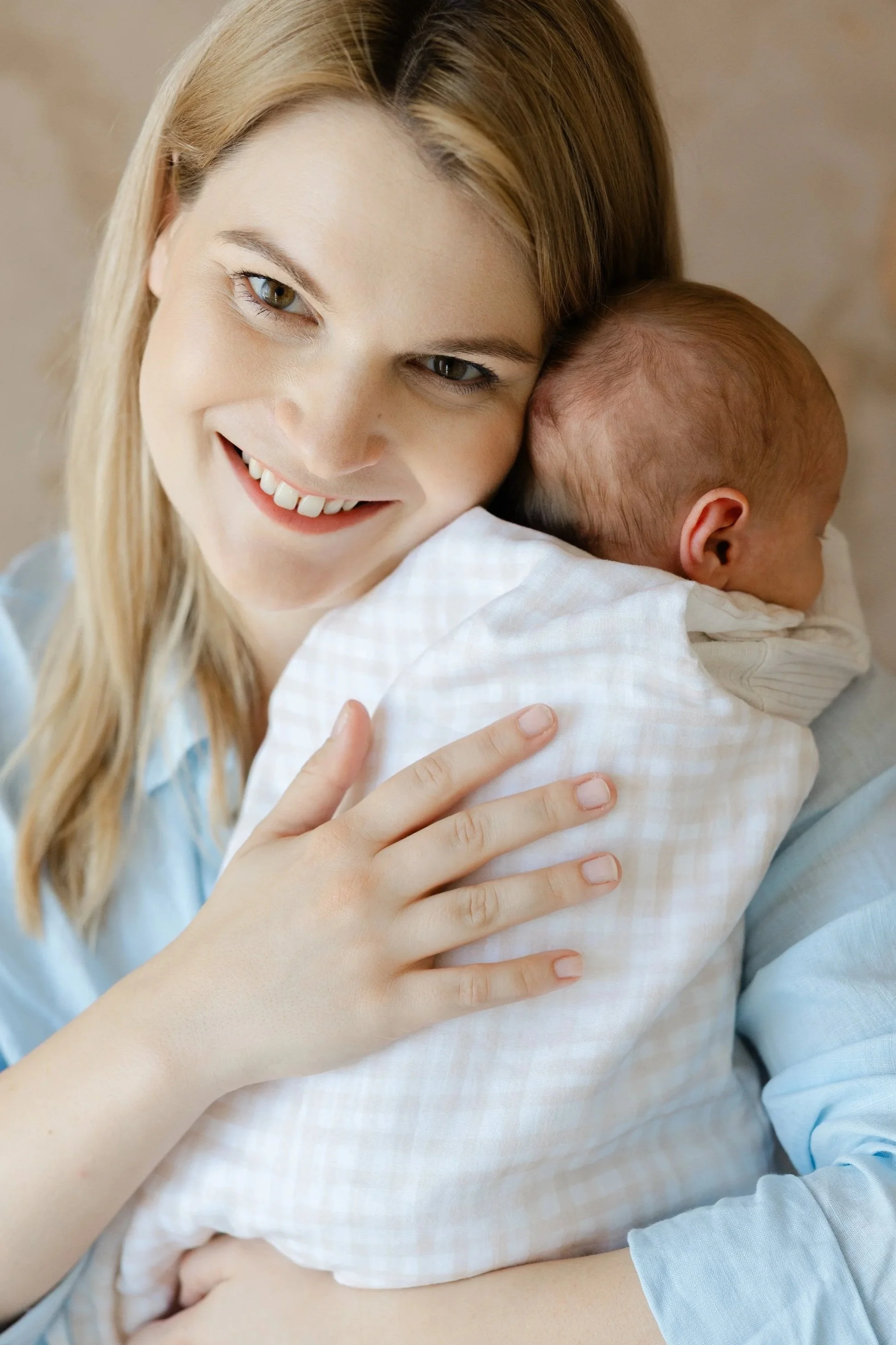 A woman with long blonde hair smiling happily while hugging a newborn baby, who has closed eyes and a tiny ear visible, dressed in a light-colored outfit.