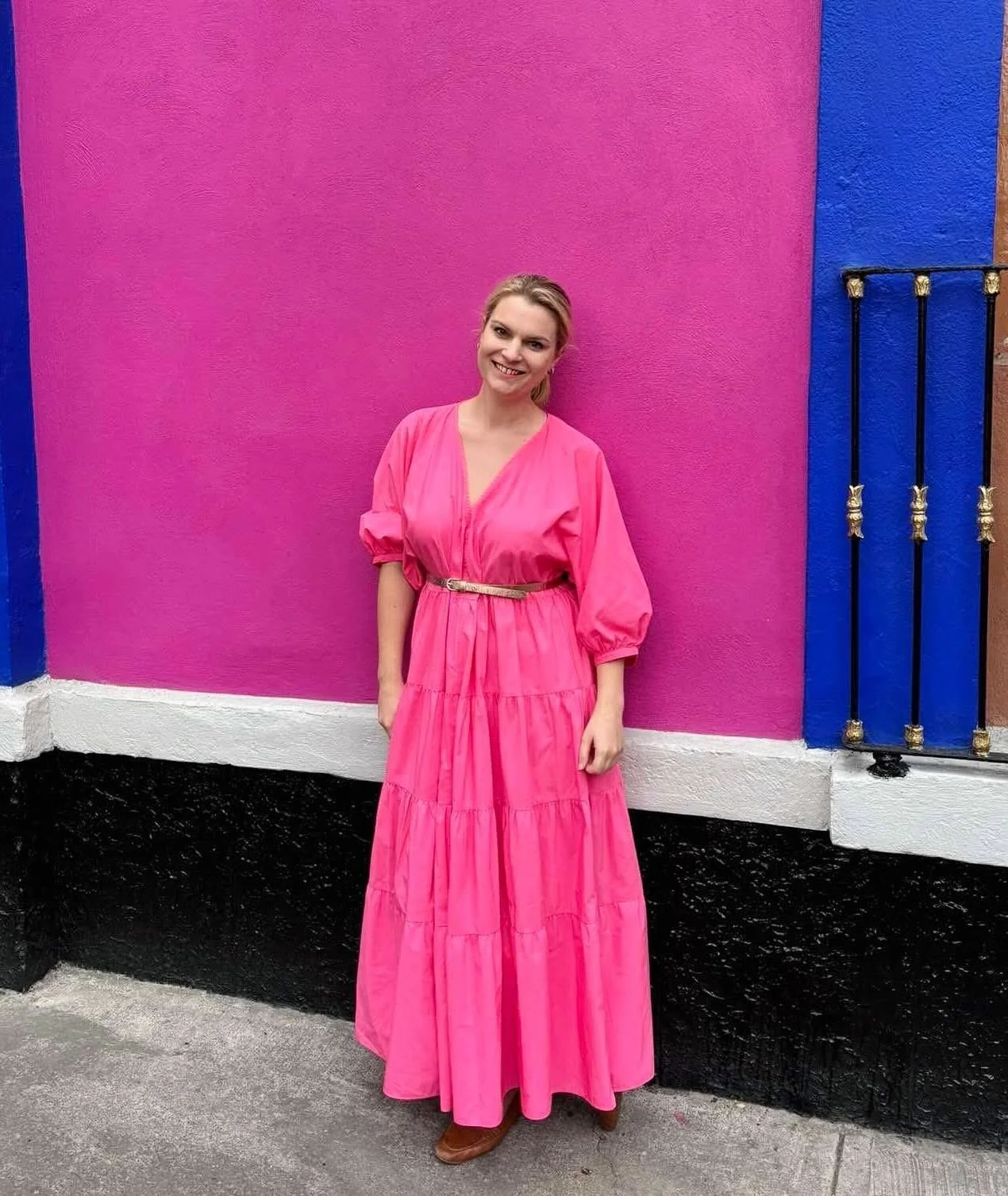 A woman in a long, bright pink dress standing in front of a colorful wall with pink, blue, and black colors and a black wrought iron gate.