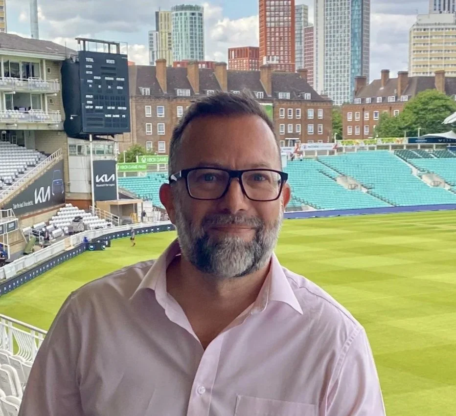 A man with glasses and a beard wearing a light pink shirt standing in a stadium with a green field, surrounded by city buildings.