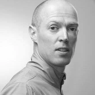 Black and white portrait of a man with a shaved head wearing a collared shirt, looking over his shoulder.