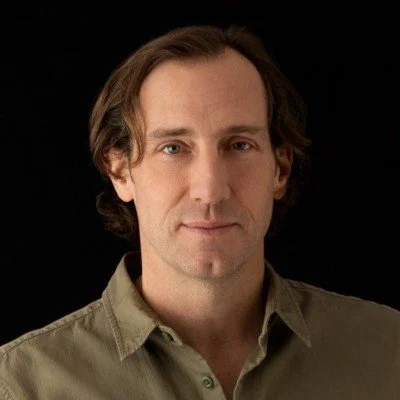 Portrait of a man with medium-length brown hair, wearing a beige button-up shirt, against a black background.