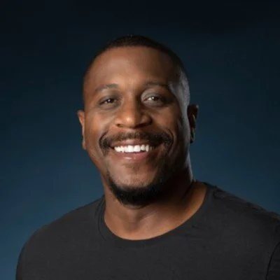 A smiling man with short hair and a goatee, wearing a black T-shirt, against a dark background.
