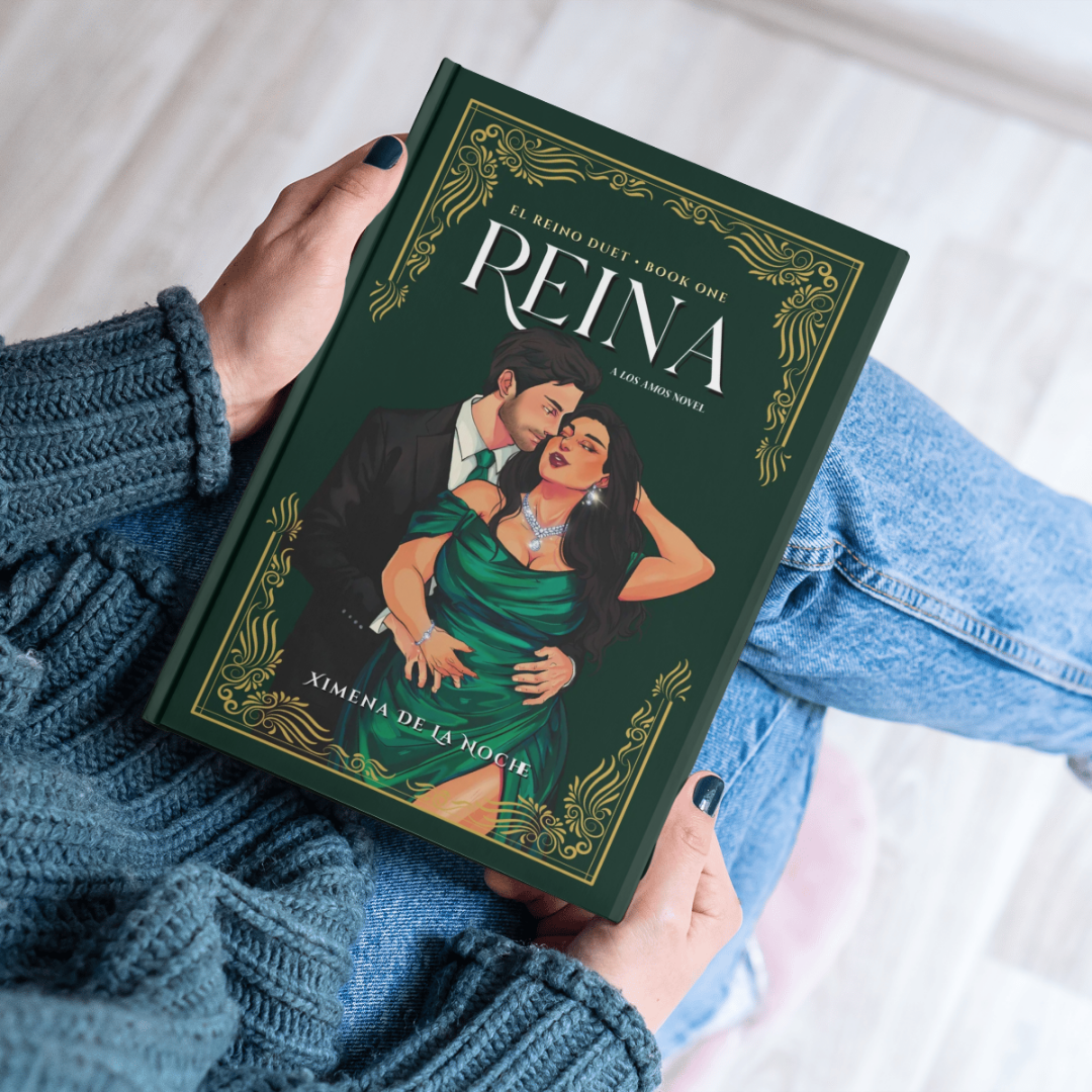 A person wearing a blue knitted sweater and jeans is sitting and holding a green romance novel titled 'Reina,' featuring an illustration of a man in a tuxedo and a woman in a green dress with jewelry, on a wooden floor.