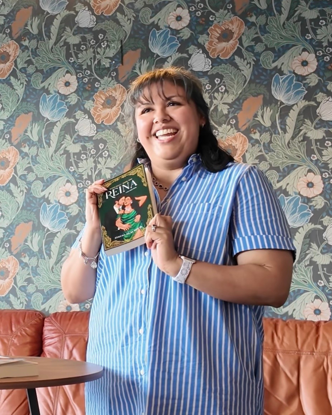 A woman smiling and holding a book titled 'Reina' in a room with floral wallpaper and a leather sofa.