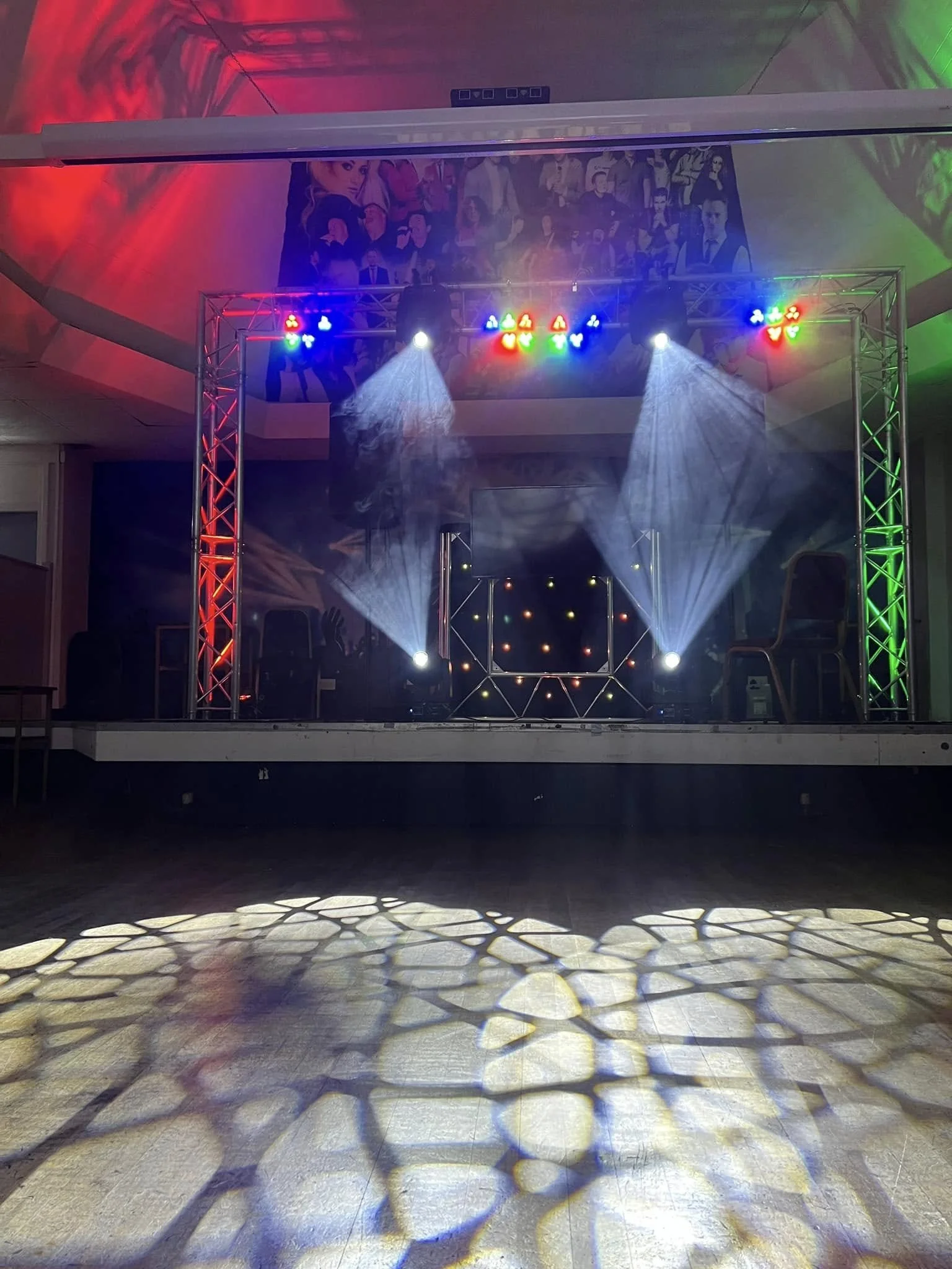 Empty dance floor with colored lights casting shadows and a stage with a backdrop of various professional photos.