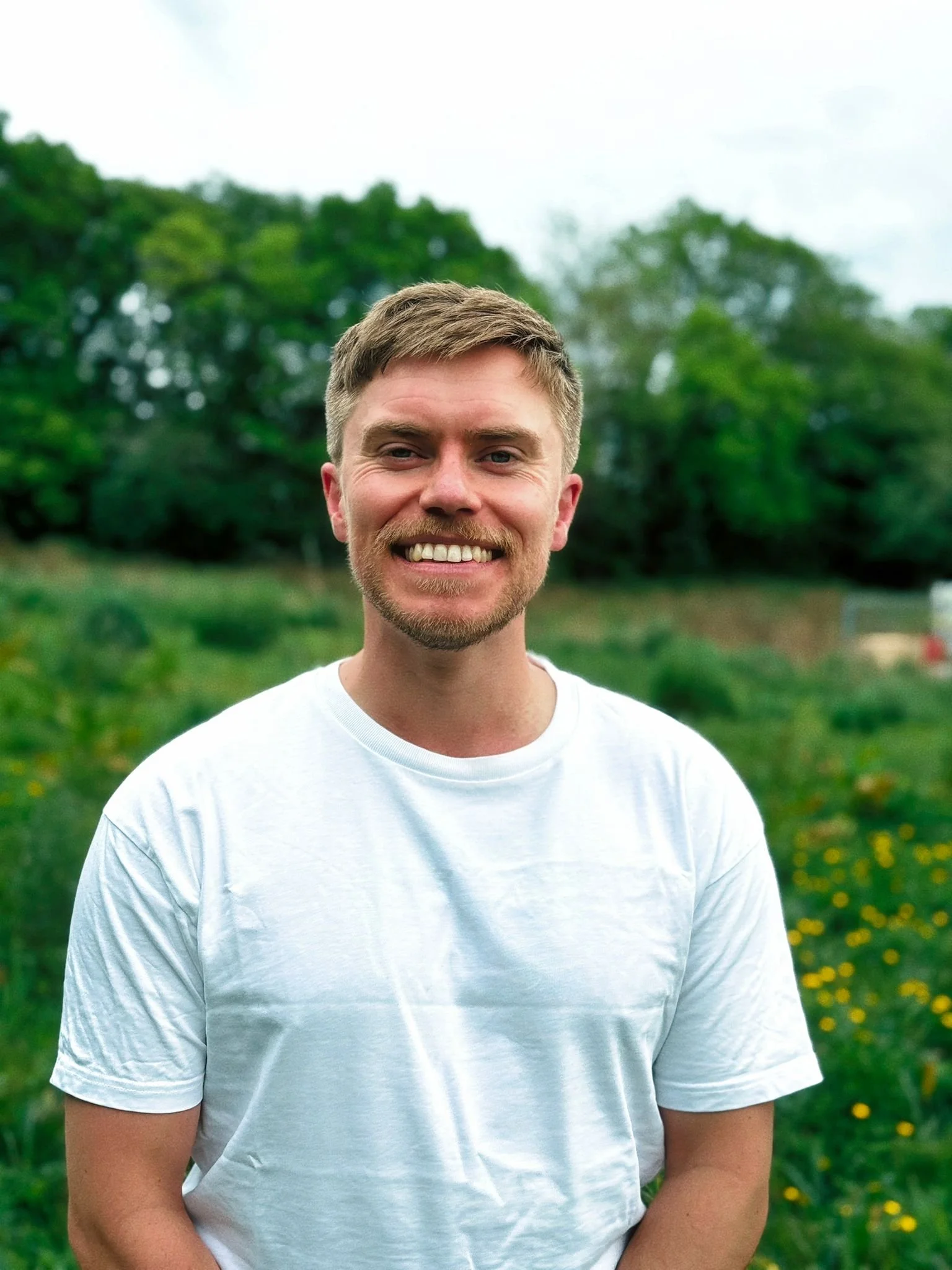 A young man with short blond hair, a beard, and a big smile, wearing a white t-shirt, standing outdoors in a green park with trees and yellow flowers, on a cloudy day.