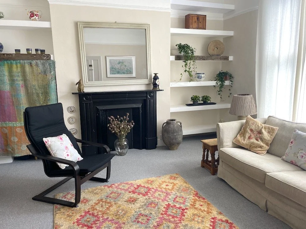 Living room with cream sofa, black armchair, patterned rug, white shelves with plants and decorative items, fireplace with mirror above, and a side table with a lamp.
