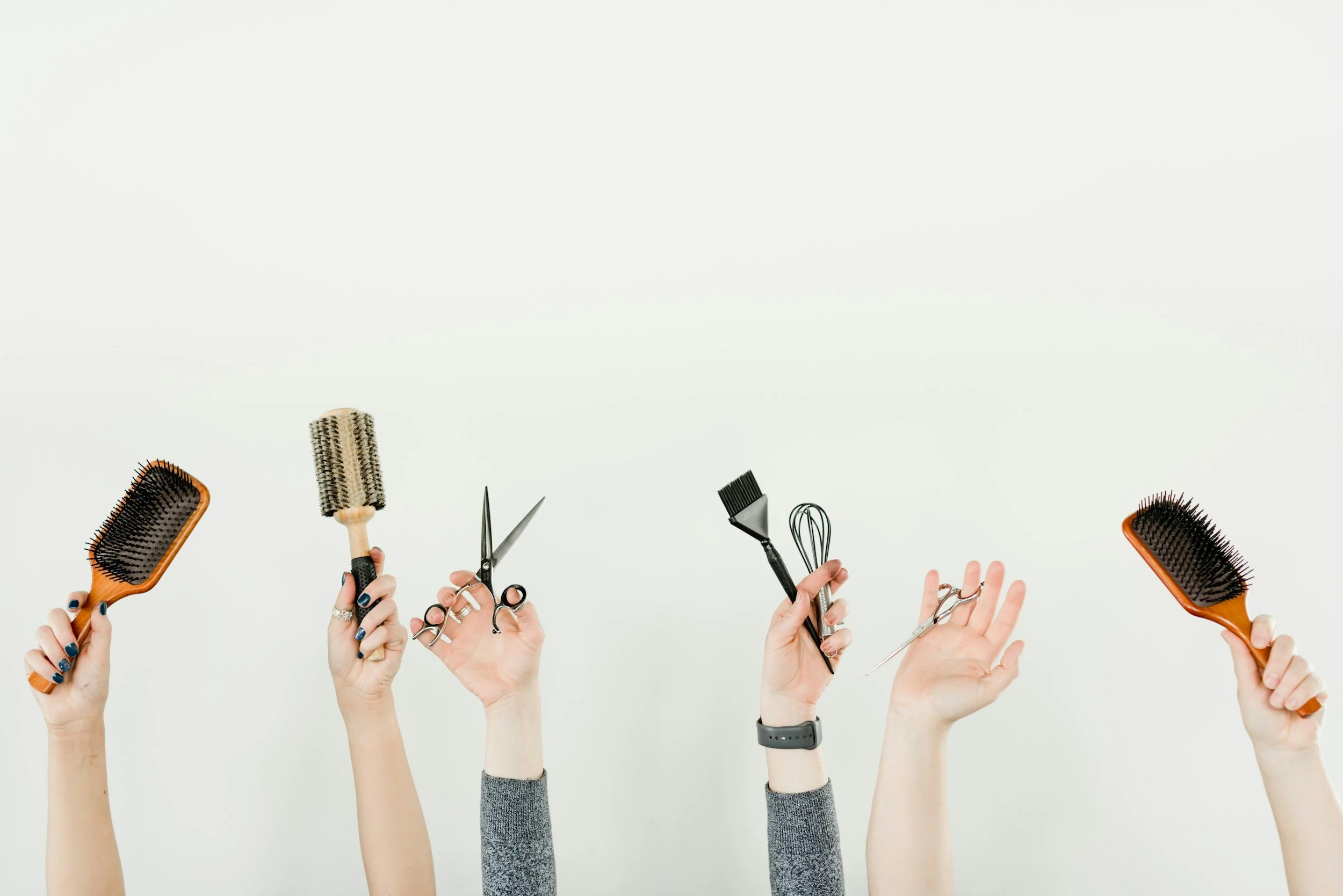 Multiple hands holding various hair styling tools, including brushes, scissors, and sprays, against a plain white background.