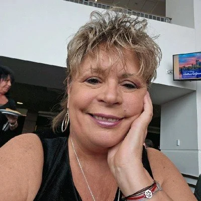 Close-up of a smiling woman with short blonde hair, wearing a black top, earrings, and a bracelet, resting her face on her hand