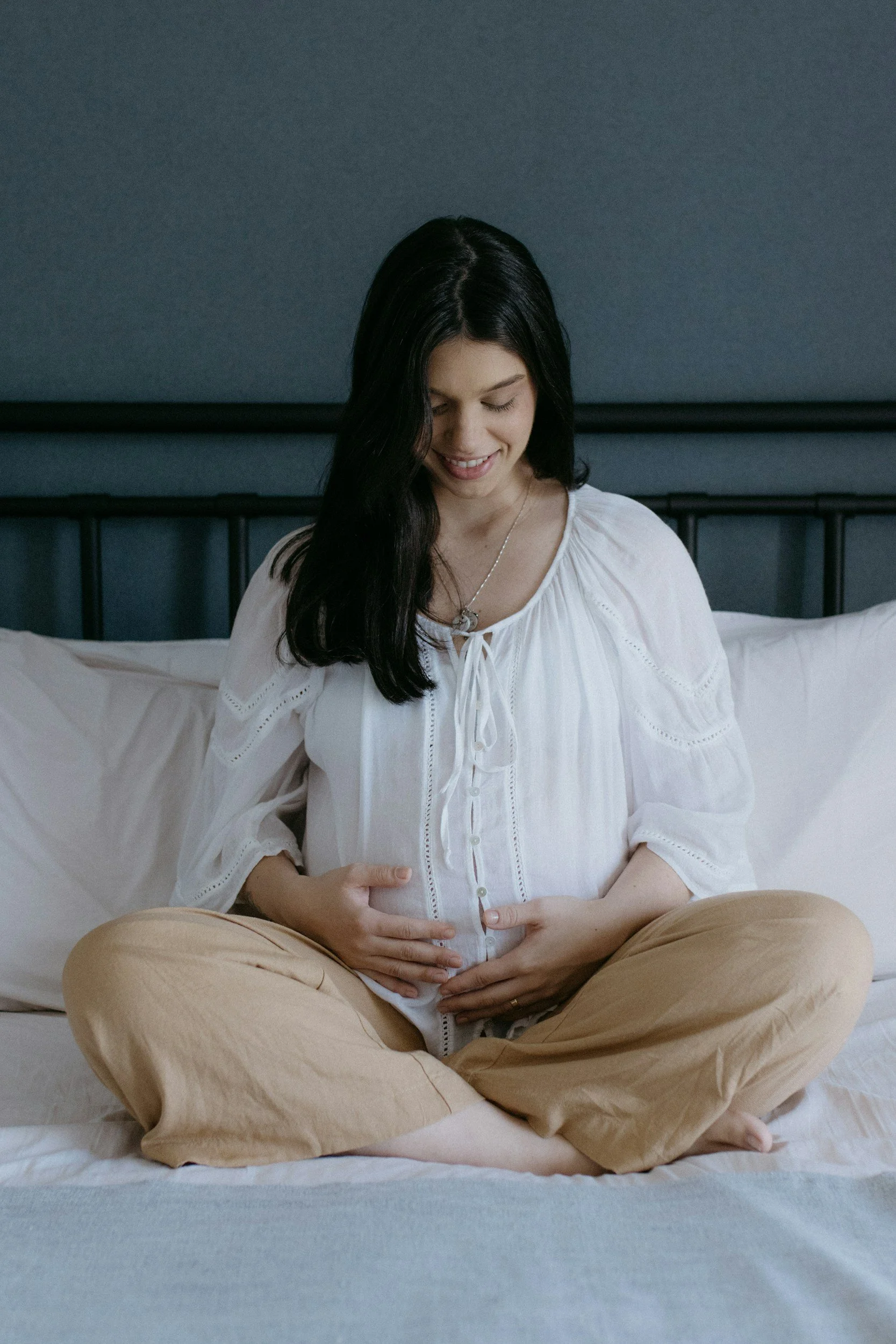 A pregnant woman with long dark hair wearing a white blouse and tan pants sitting cross-legged on a bed, smiling and holding her belly with both hands. The background includes a dark headboard and gray wall.