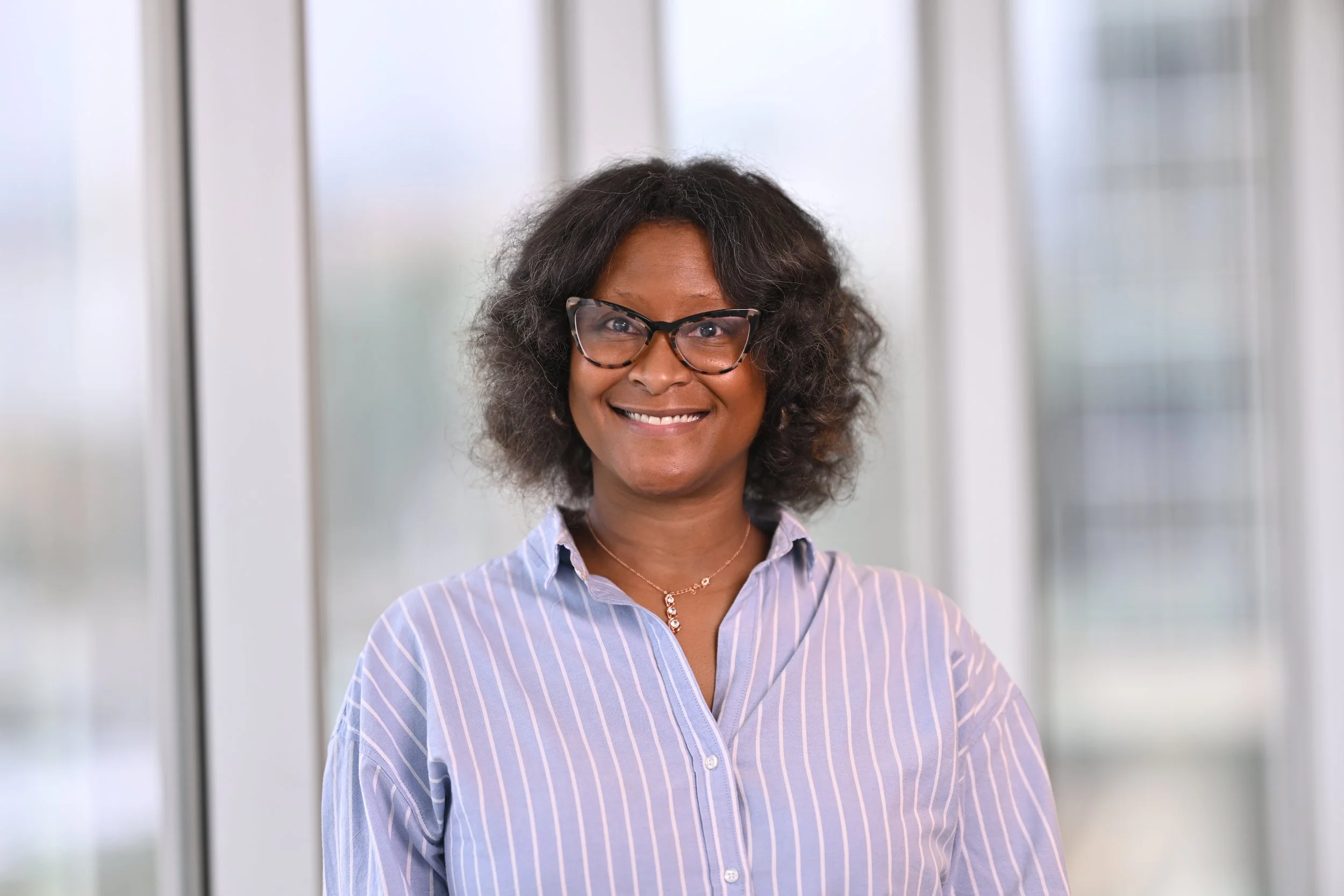 Smiling African American woman with glasses and curly hair wearing a light blue striped shirt indoors.