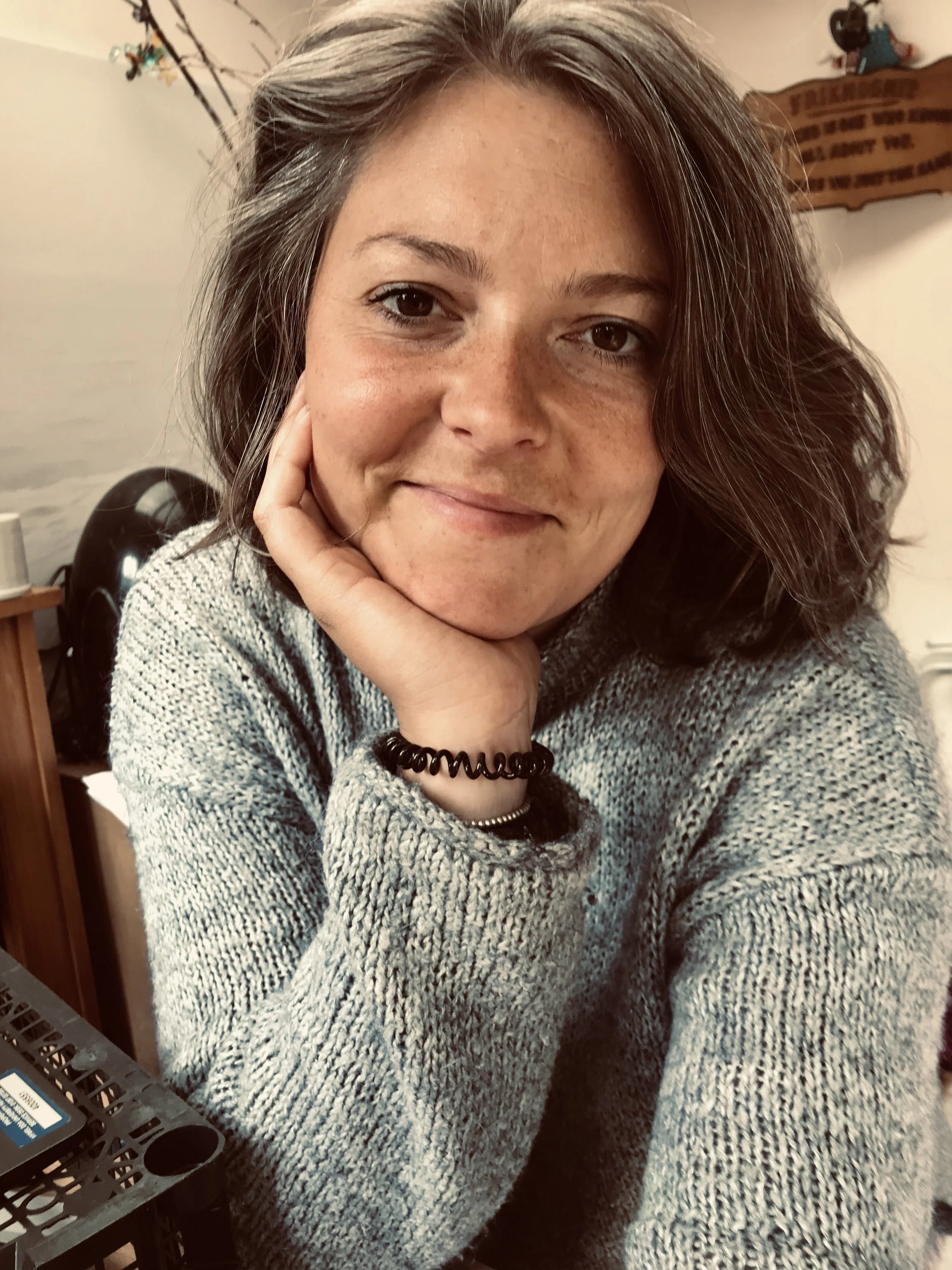 A woman with shoulder-length wavy gray hair, wearing a gray knitted sweater and black elastic hair tie, resting her face on her hand and smiling softly.
