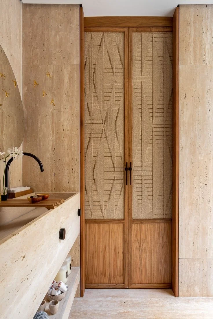 A wooden closet with textured sliding doors featuring a leafy pattern, situated next to a beige stone bathroom wall with a countertop and black faucet.