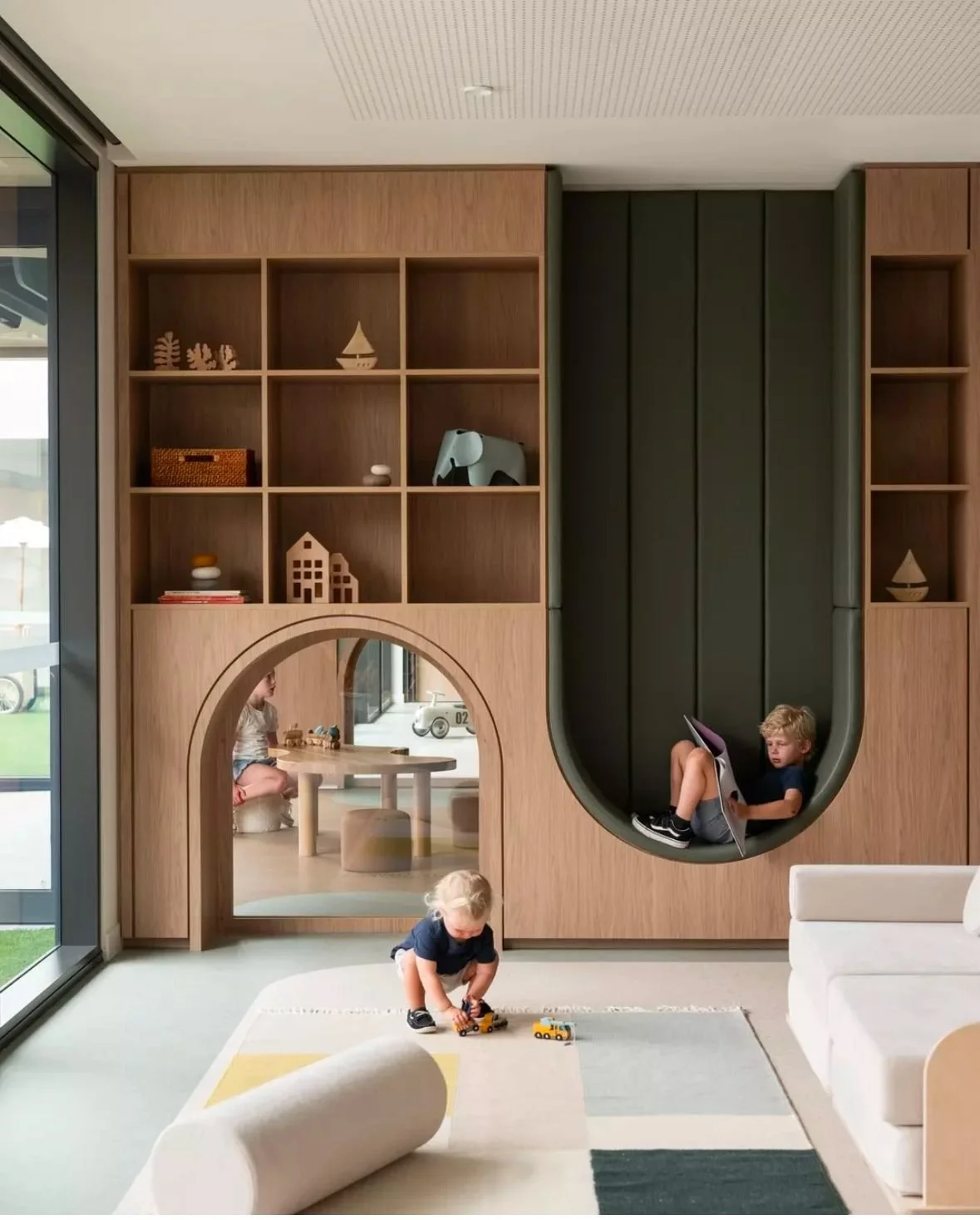 Children playing in a modern, stylish interior with built-in shelving, a small play area, and a large window bringing in natural light.