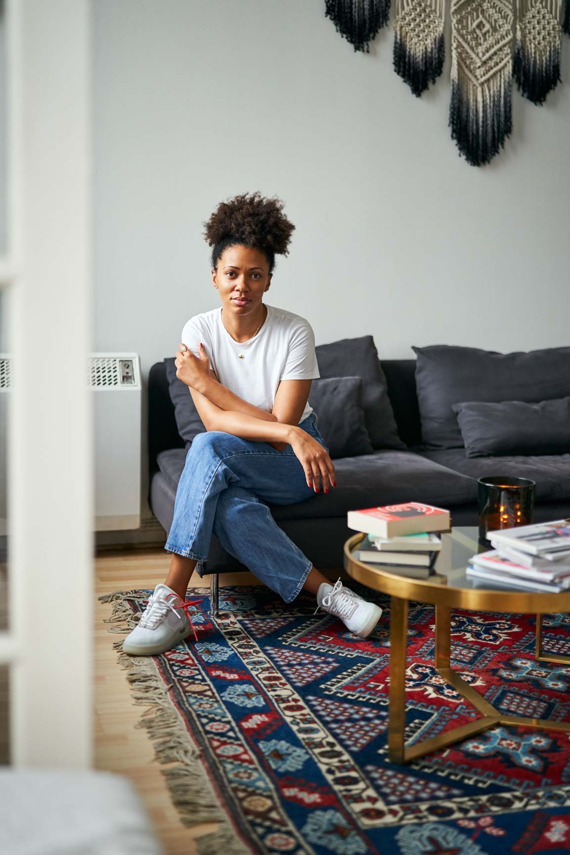 Maïna Cissé sitting on a dark gray couch with her arms crossed, in a living room with a colorful patterned rug and a round coffee table with books, a candle, and a glass. Wall hanging decor is visible behind her.